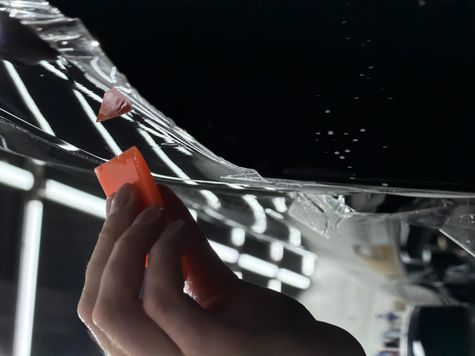 The image shows a person's hand using an orange squeegee to apply a transparent film to a glossy black surface. Water droplets are visible between the film and the surface. The background features a blurred, out-of-focus environment with linear light fixtures, suggesting an indoor setting like a garage or workshop. The specific location in Krasnoyarsk, Russia, cannot be determined from the image.