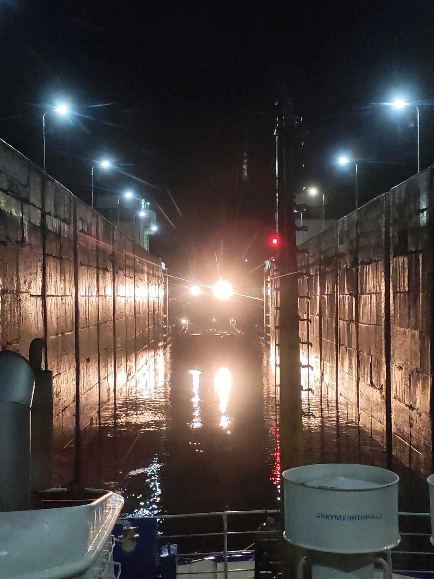 The image depicts a nighttime scene inside a ship lock, likely in Markovo, Russia. The lock walls are tall and made of concrete blocks, illuminated by numerous streetlights and the bright headlights of an approaching vessel. The water in the lock reflects the lights, creating shimmering patterns. In the foreground, part of a white boat or vessel is visible, including a railing and a cylindrical metal container with Russian text that reads "АККУМУЛЯТОРНАЯ" (AKKUMULYATORNAYA), meaning "accumulator" or "battery". A red light is visible on a pole to the right of the frame. The scene appears calm and operational, with no people visible.