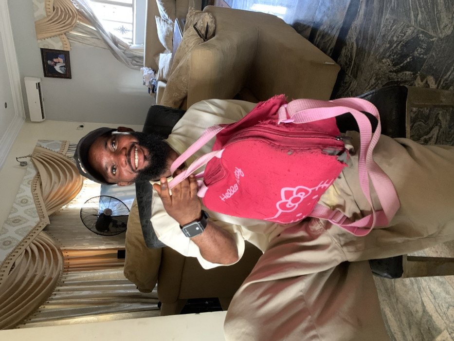 A man with a beard and baseball cap smiles, holding a torn pink "Hello Kitty" backpack. He wears an Apple watch and a white earbud. He is dressed in a cream-colored outfit and sits in a chair. A beige couch is behind him. Heavy beige drapes frame a window. An air conditioning unit is visible near the ceiling. A framed photo hangs on the wall. A fan is visible behind the man.