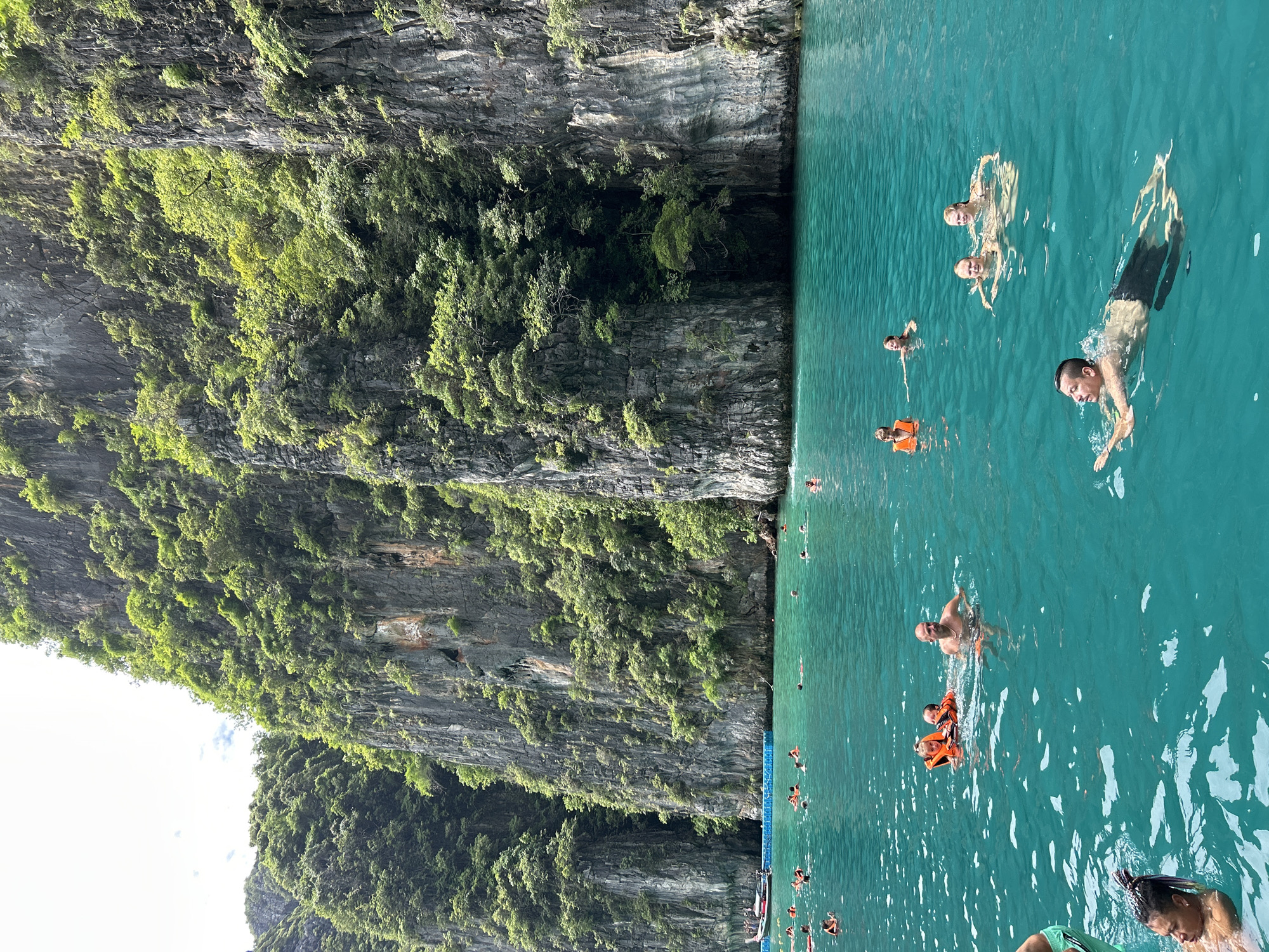 This image depicts a scenic view of a group of people swimming and enjoying the turquoise waters in Haad Yao, Thailand. Towering, lush green cliffs flank the water, creating a dramatic backdrop. Several individuals are visible in the water, some wearing orange life vests, suggesting a family or tour group. People are engaged in swimming, with some appearing to relax while others are actively moving through the water. In the background, a long-tail boat is moored near a floating blue platform, and a few other boats are visible further out. The water is clear and inviting, with sunlight reflecting off its surface, creating a tranquil and picturesque scene.