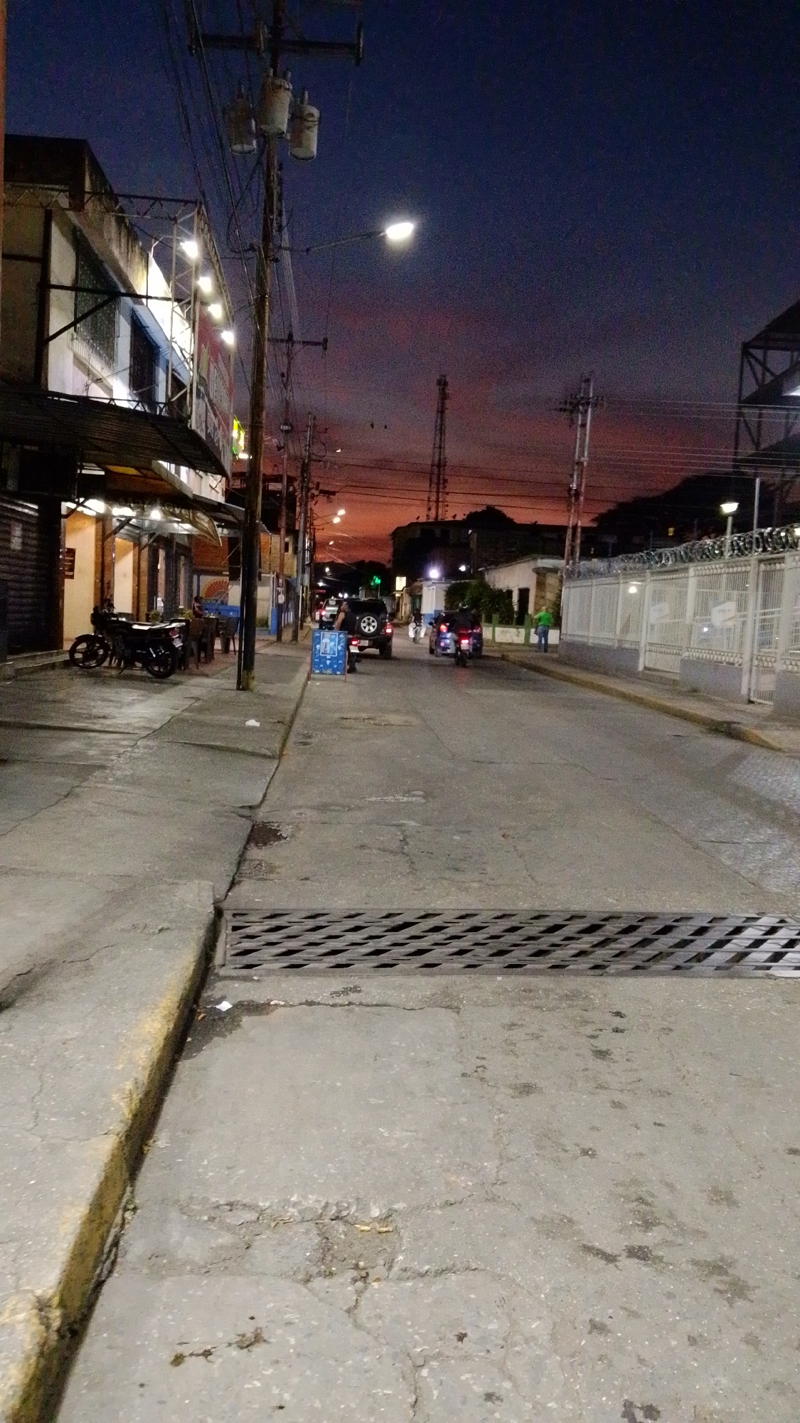 The image depicts a street scene in Tinaquillo, Venezuela at dusk. The sky exhibits a gradient from deep blue to orange-red near the horizon. The street is paved and has a storm drain with a metal grate in the foreground. A sidewalk runs along the left side of the street, lined with commercial buildings and a restaurant with outdoor seating. Several motorcycles are parked near the restaurant. Further down the street, a dark SUV is parked near a blue sign or object. A motorcycle and pedestrian are visible moving in the distance. The buildings on the right side of the street include a tall white fence topped with barbed wire. Utility poles with power lines are prominent on both sides of the street. Streetlights illuminate the scene.