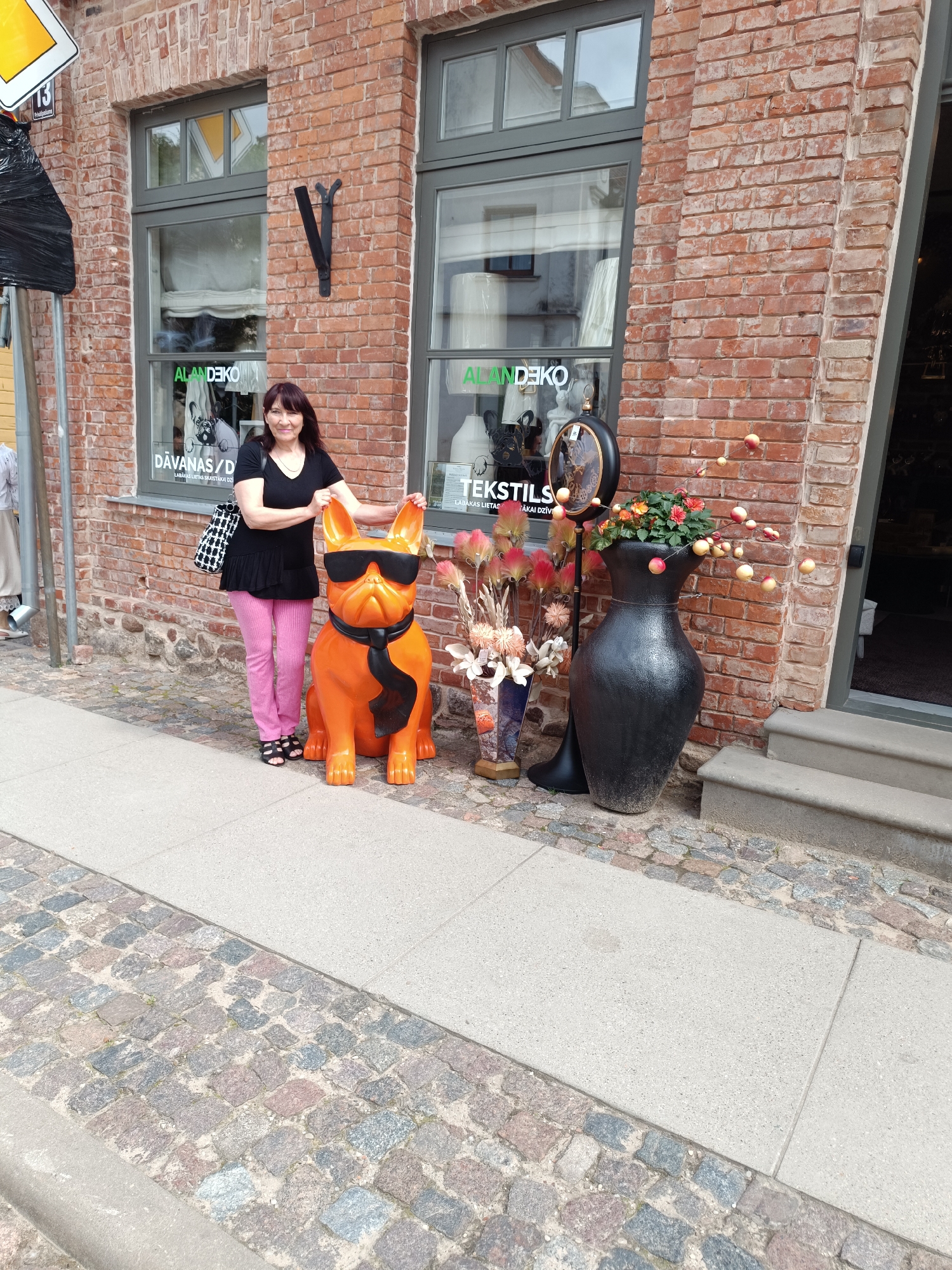 A woman stands on a cobblestone sidewalk in front of a brick building in Kuldīga, Latvia. She is wearing a black top, pink pants, and black sandals. She has dark hair and is smiling. Beside her is a large, orange statue of a French bulldog wearing sunglasses and a black tie. In front of the building are two large vases, one black and one decorative, with flowers and artificial apples. The building has large windows with signage that reads "ALANDEKO". A sign with the number 13 is visible on the left side of the image.