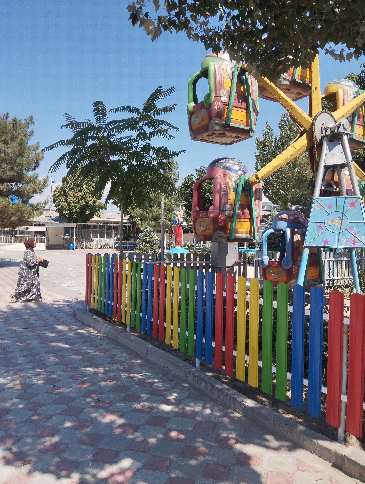 A Ferris wheel with colorful gondolas stands in the foreground of an outdoor amusement park. The Ferris wheel is yellow and grey, with red, yellow, blue, and green gondolas. A woman in a patterned dress and headscarf walks on a paved path in front of a colorful fence. The fence is made of vertical wooden slats painted red, yellow, green, and blue. In the background, there is a building with large windows and trees. The sky is clear and blue. The scene appears to be in Tokmok City, Kyrgyzstan, as indicated by the context.