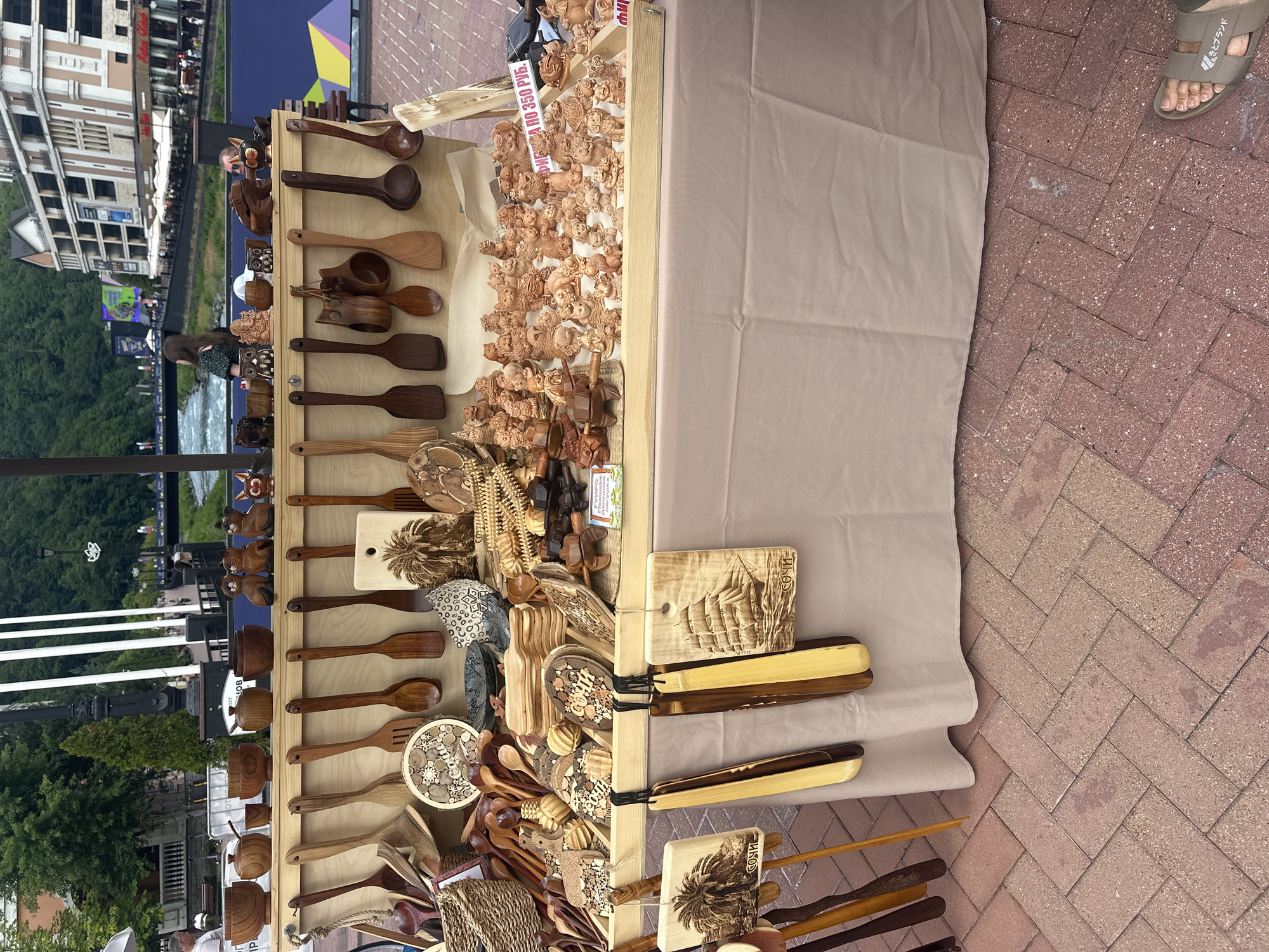 The image shows an outdoor market stall in Estosadok, Russia, with various wooden crafts and kitchenware for sale. The stall is set up on a paved walkway with a river and buildings in the background, suggesting a resort or tourist area. The stall displays a collection of hand-carved wooden animals, bowls, spoons, and decorative items. The items are arranged on shelves and hanging from the stall. Some of the items have Cyrillic inscriptions, indicating they are likely souvenirs. A price tag in Russian currency is visible on one of the items. The overall scene depicts a typical market environment in a tourist destination.