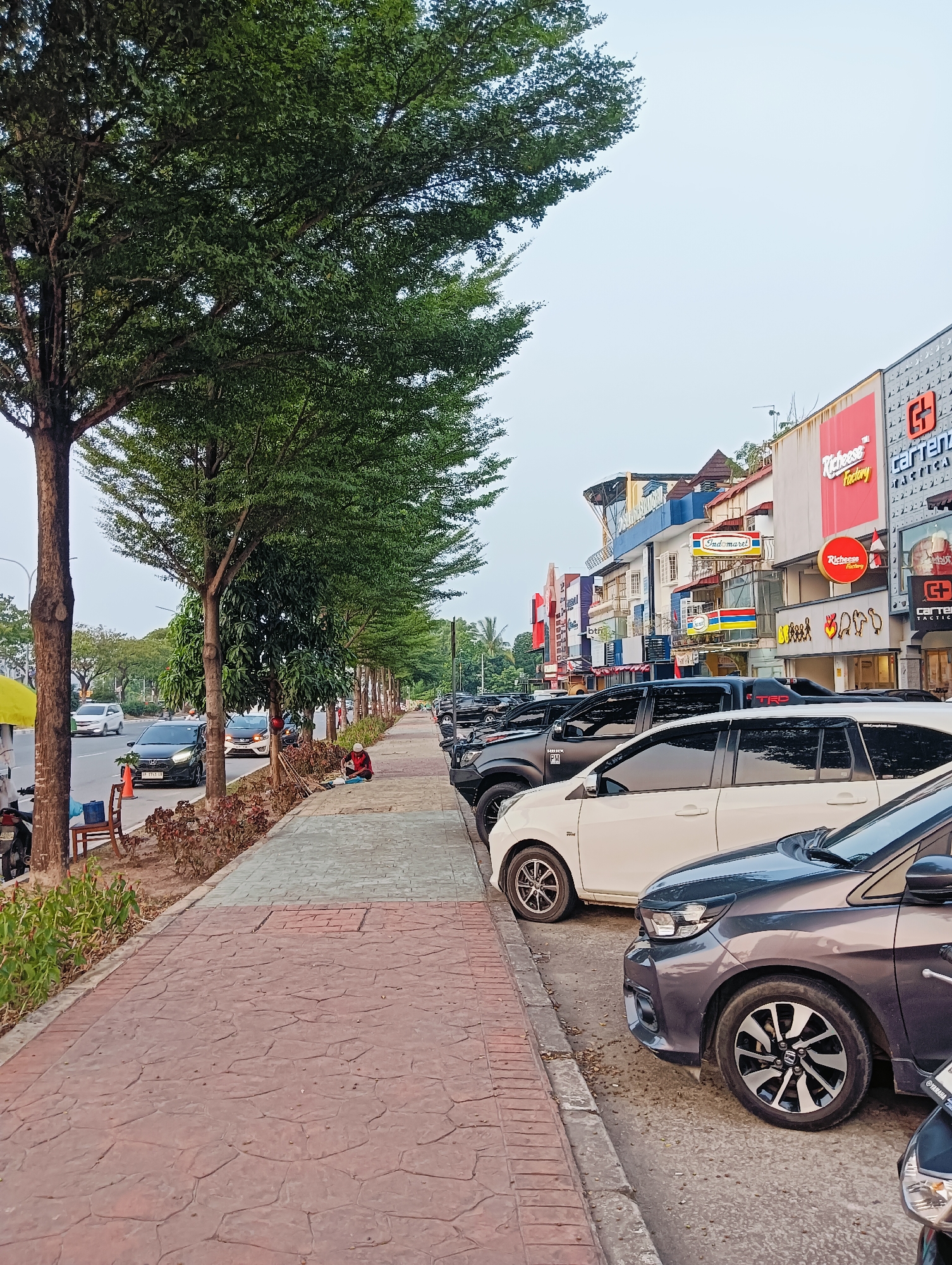 The image depicts a street scene in Batam, Indonesia, characterized by a row of parked cars and shops lining the right side of the road. A pedestrian walkway, paved with textured concrete, runs alongside the street. Trees with dense green foliage are planted along the walkway, creating a shaded path. A person is seated on the walkway near some plants. On the opposite side of the street, vehicles are in motion. The shops have prominent signage, with "Richeese Factory" and "Indomaret" being clearly visible. The sky is overcast, suggesting either early morning or late afternoon lighting conditions. The overall impression is that of a typical urban commercial area.