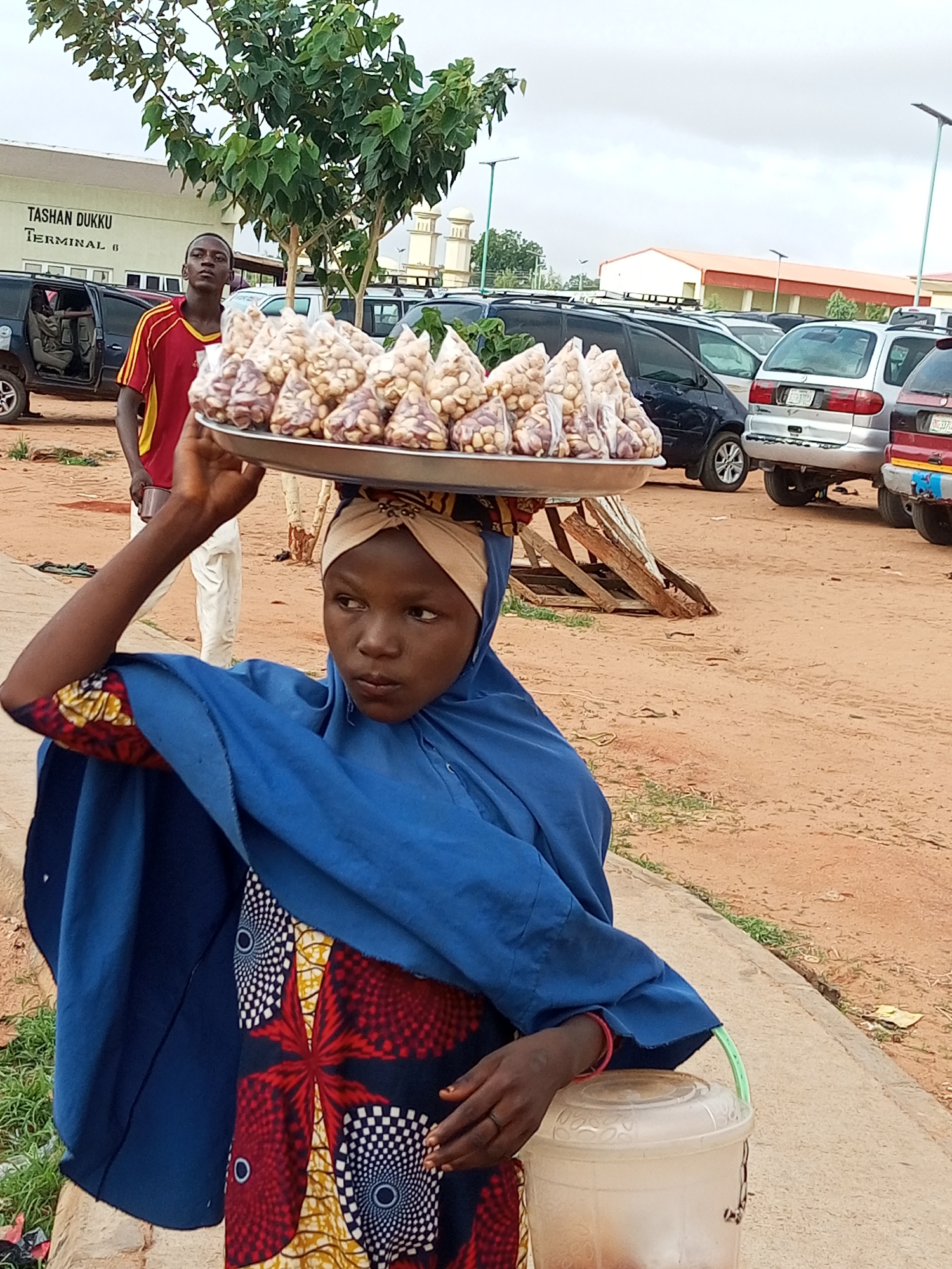 A young Nigerian girl, possibly in her early teens, carries a large metal tray laden with small, clear plastic bags filled with what appears to be nuts or dried snacks on her head. She is wearing a vibrant, patterned cloth skirt and a blue headscarf and outer garment. Her head is slightly tilted, and her gaze is directed to her left. To her left, a young man in a red and yellow striped shirt stands with his arms crossed, looking towards the girl.
The background suggests an outdoor market or transport hub. A building with "TASHAN DUKKU TERMINAL 8" signage is visible on the left, indicating the location is Tashan Dukku Terminal 8 in the city of Gombe, Nigeria. Several vehicles, including SUVs and vans, are parked behind the individuals. The ground is covered in reddish-brown dirt, typical of arid or semi-arid regions. A concrete pathway runs in the foreground, where the girl is standing. The sky is overcast. The overall scene depicts a common sight in Nigerian marketplaces, where vendors, often young people, carry goods to sell. The girl's posture and the tray balanced on her head are characteristic of traditional methods of transporting merchandise.