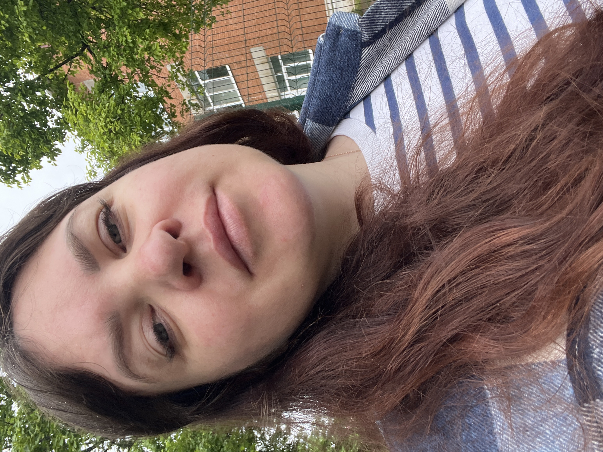 A close-up portrait, oriented sideways, shows a young adult individual, likely female, looking directly into the camera. She has fair skin, dark, possibly brown eyes, and full lips. Her long, wavy hair, which appears to be a shade of brown with reddish undertones, frames her face and extends across the frame. Her expression is calm and neutral, with a hint of a slight smile.
She is wearing a white t-shirt with thin vertical blue stripes. A blue and white plaid or striped jacket, with a denim-like texture, is draped over her right shoulder. A thin gold chain necklace is visible around her neck.
The background, viewed from a low angle, features a lush green tree canopy on the left side, indicating an outdoor setting. Behind the foliage and to the right, a red brick building with several windows is visible, partially obscured by a dark metal fence or grating. The sky appears bright white and overcast, suggesting it is daytime with diffused lighting. The scene conveys a relaxed moment in an urban or suburban outdoor environment, located in Noviy, Russia. There is no visible text in the image.
