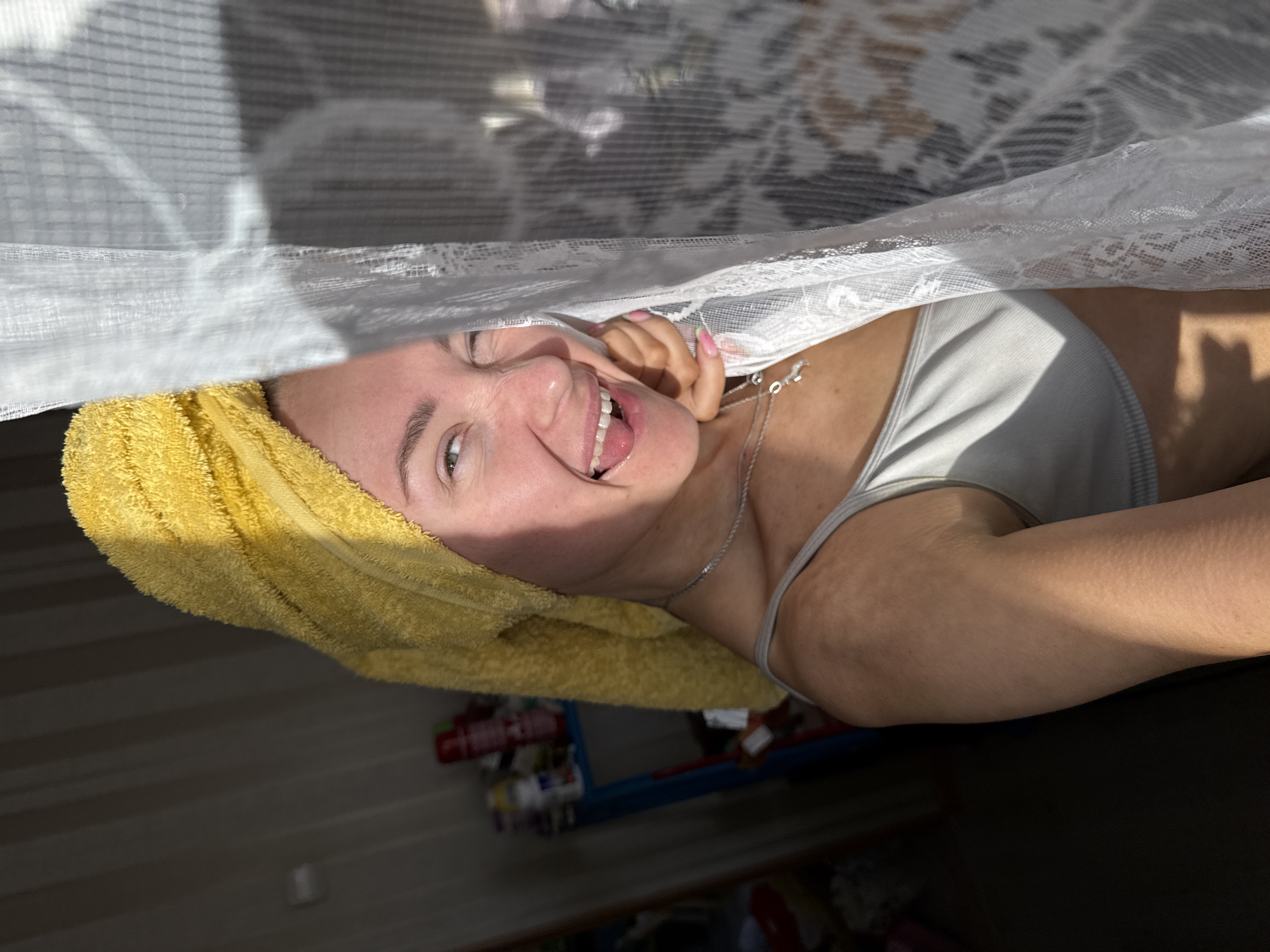 The image captures a bright and candid indoor moment featuring a young woman playfully peeking from behind a white, sheer curtain, possibly made of lace. She is smiling broadly with an open mouth, revealing her teeth and tongue, and her eyes convey happiness and cheerfulness. A vibrant yellow terry cloth towel is wrapped around her head, indicating she may have recently washed her hair or showered. She is wearing a light grey tank top and a delicate silver chain necklace with a small, shiny charm. Her hand, adorned with light pink or French-tipped fingernails, is visible near her face, appearing to hold the curtain aside.
The scene is well-lit by natural sunlight, streaming from the left side of the frame, which casts distinct shadows across her face and arm, and creates geometric light patterns on the background fabric or wall. The background beyond the curtain is dark and out of focus, suggesting an indoor room with indistinct elements like shelves holding various items and a blurry object with a blue frame. The overall atmosphere is joyful and relaxed, set within a domestic environment, likely in Chaikovsky, Russia, during a sunny day. No visible text is present in the image.
