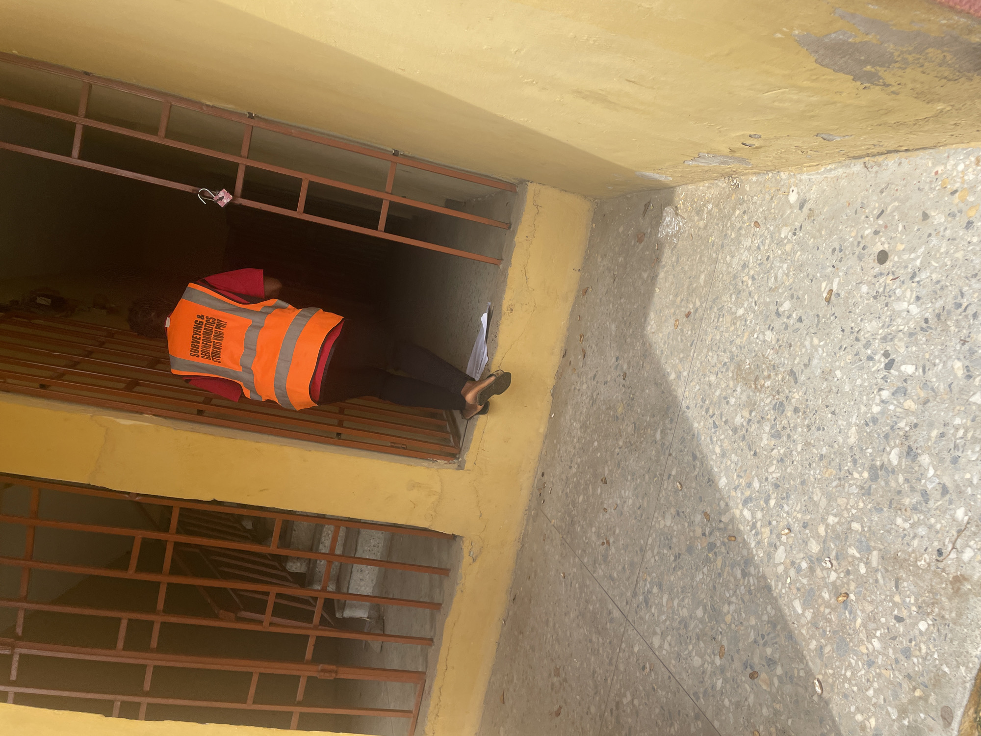 A person wearing an orange reflective vest and black pants stands at the entrance to a dark corridor with a locked metal gate. The vest has text on the back that reads "SURVEYING & GEOINFORMATICS STUDENTS KOGI POLY." The person is facing away from the camera and appears to be stepping over some papers on the ground. The building exterior is painted yellow, and the ground is a speckled concrete material. The scene is likely outdoors in a public space. The context suggests this is in Lokoja, Nigeria, given the text on the vest referencing "KOGI POLY," which is located in Lokoja.