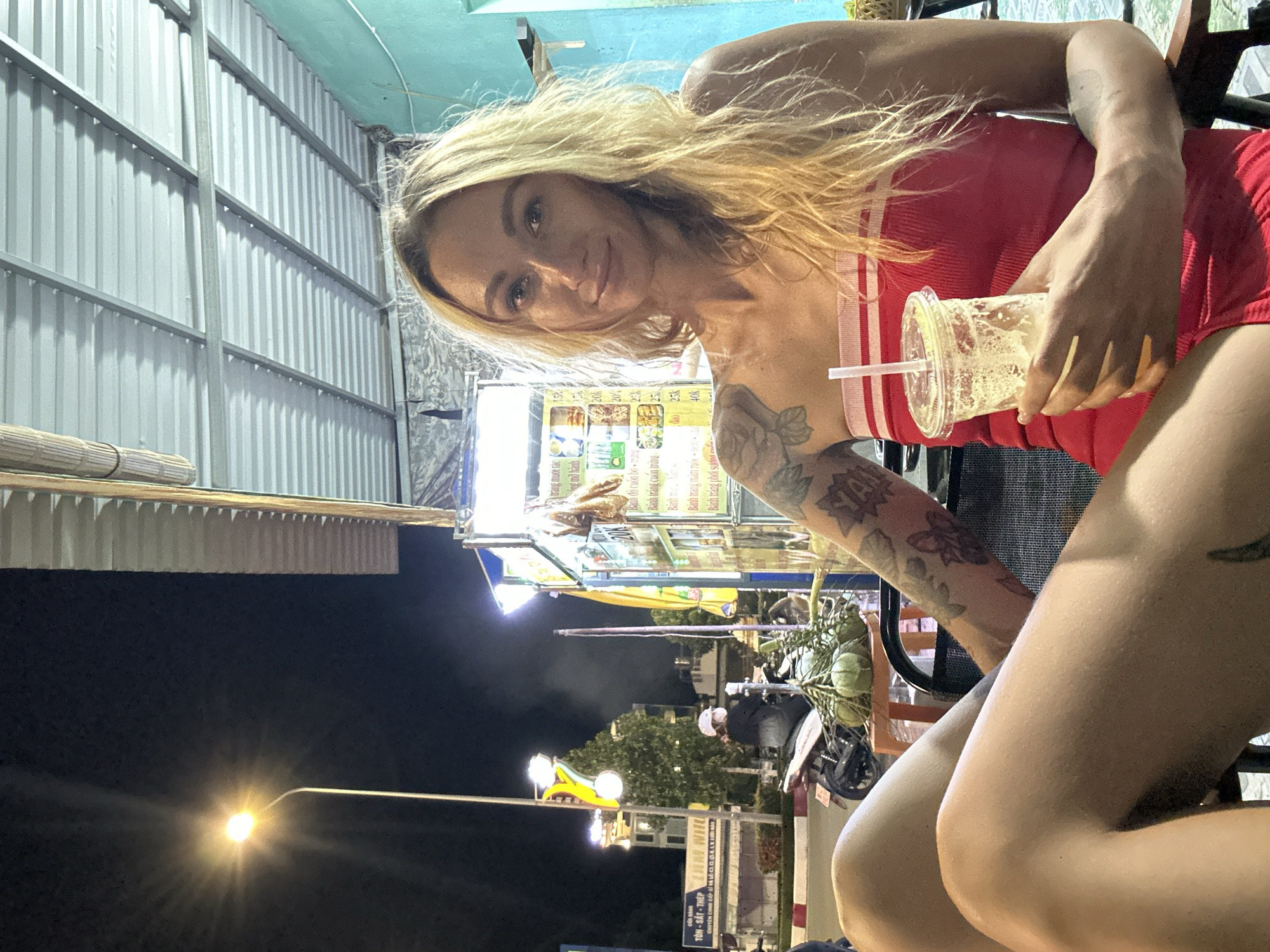 A young Caucasian woman with blonde hair is seated at an outdoor establishment at night, holding a plastic cup with a straw and an iced beverage. She is wearing a red strapless top with white stripes. Her left arm is visible, and appears to have tattoos. The background shows a street scene with a lamppost, a food stall with illuminated signs displaying text in Vietnamese, and a person on a motorbike in the distance. The establishment has a corrugated metal roof. The scene is consistent with a street-side eatery or market in Phu Thuy Ward, Vietnam.
