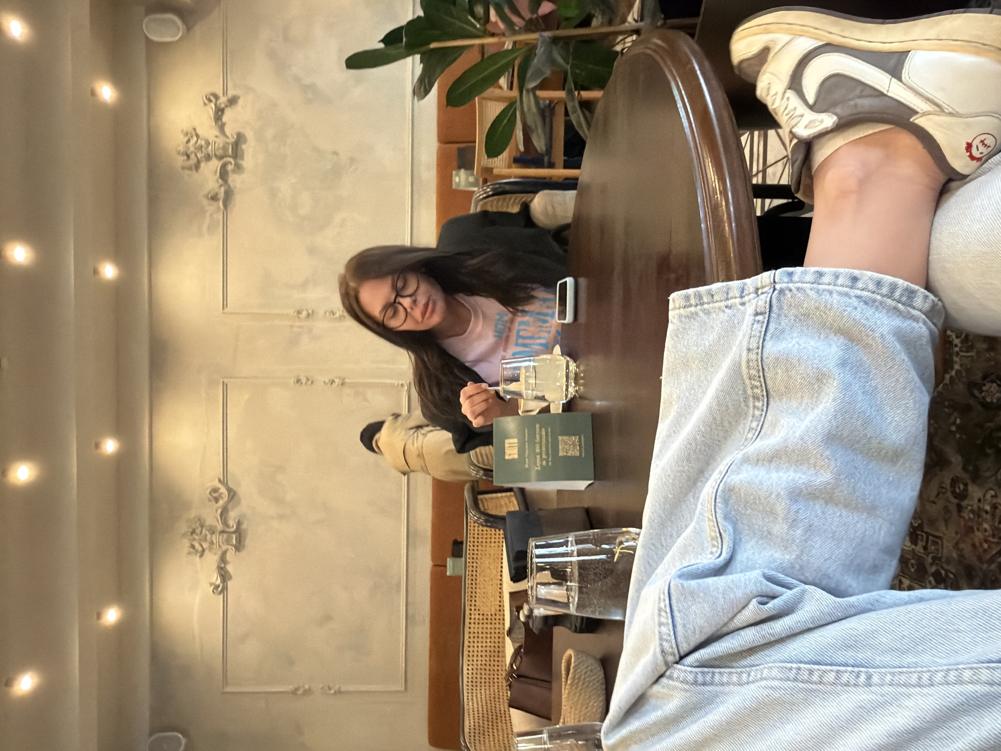 The image captures a casual scene indoors, likely a restaurant or cafe in Krasnoyarsk, Russia. A woman with glasses is seated at a dark wooden table, wearing a pink t-shirt and a dark jacket. She seems to be looking at something on the table, possibly a drink or a menu. An individual's legs are visible in the foreground, clad in light blue denim jeans and wearing grey and white sneakers. A green card with the name "SKITO" is on the table, alongside multiple glasses. The interior features textured walls with decorative moldings and multiple lights. A plant is partially visible in the background, contributing to the restaurant's ambiance. Other people are present in the background.