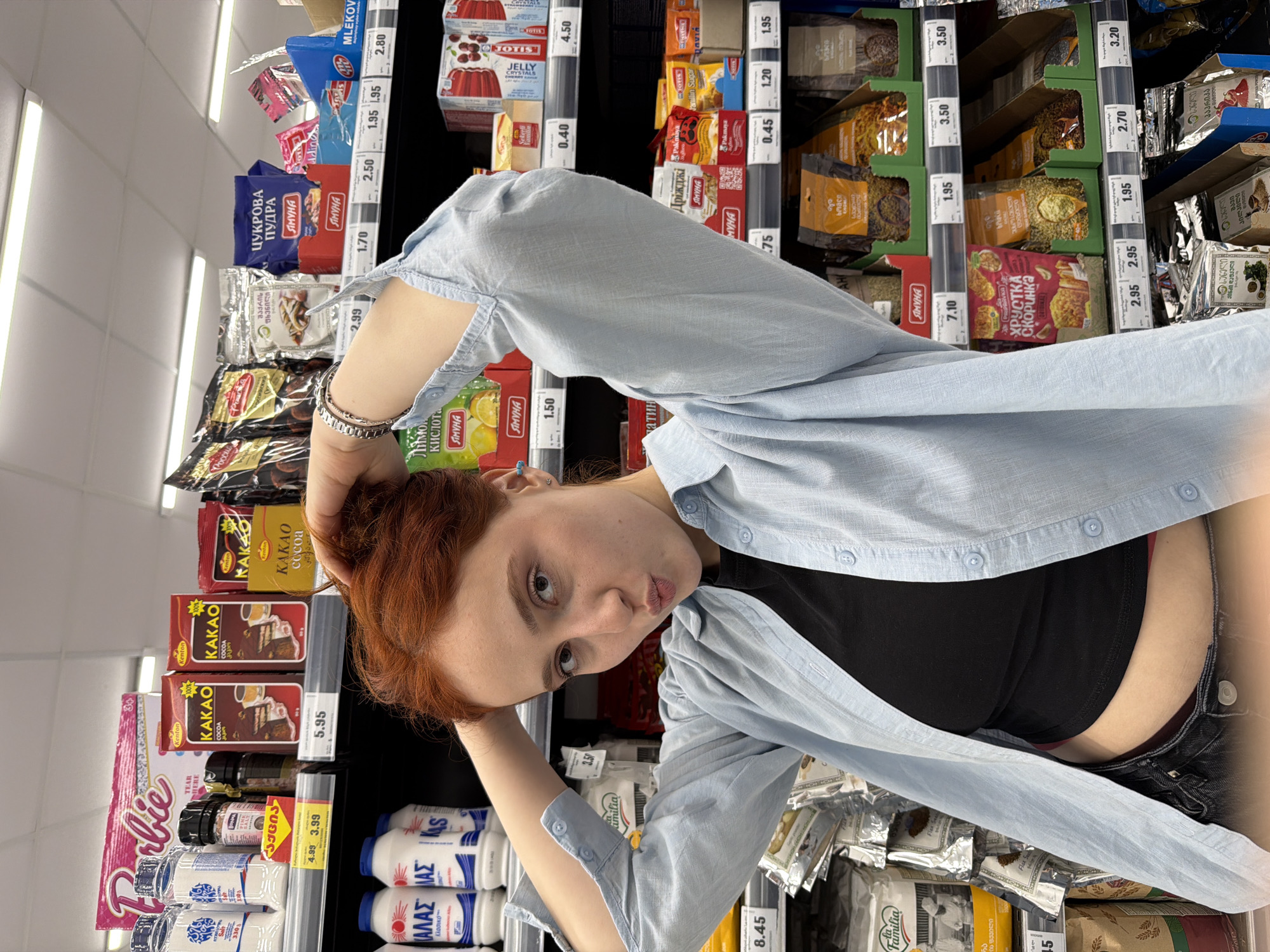 A young woman with red hair and light skin is captured in a playful pose within a grocery store aisle in Tbilisi, Georgia. She wears a light blue long-sleeved shirt over a black top, paired with dark bottoms. Her head is tilted, and she looks towards the camera with a slight pout, her left hand resting behind her head/neck and her right hand near her hair. A metallic bracelet adorns her left wrist.
The setting is a brightly lit supermarket aisle, with fluorescent strip lights visible on the ceiling. Numerous shelves line the background, packed with a wide array of packaged food items, predominantly groceries and baking ingredients. Price tags with various numerical values are visible throughout the shelves.
Visible text on product packaging includes "Barbie," "Kenton KAKAO cocoa," "JOTIS JELLY CRYSTALS," "MLEKOV," and items from the "SIMYHA" brand such as "ЦУКРОВА ПУДРА" (Sugar Powder) and "Лимон Кислота" (Citric Acid). Other discernible brands and products include "Pakmaya" (yeast), "ХРУСТКА Скоринка" (Crispy Crust), and "Familia." Bottles labeled with "ΚΑΛΑΣ" (Kalas) in Greek script are also present. Additionally, Georgian script, such as "პაპრიკა" (Paprika), is visible on some labels, reflecting the Tbilisi, Georgia location. The overall scene suggests a casual, perhaps humorous, moment during a shopping trip.