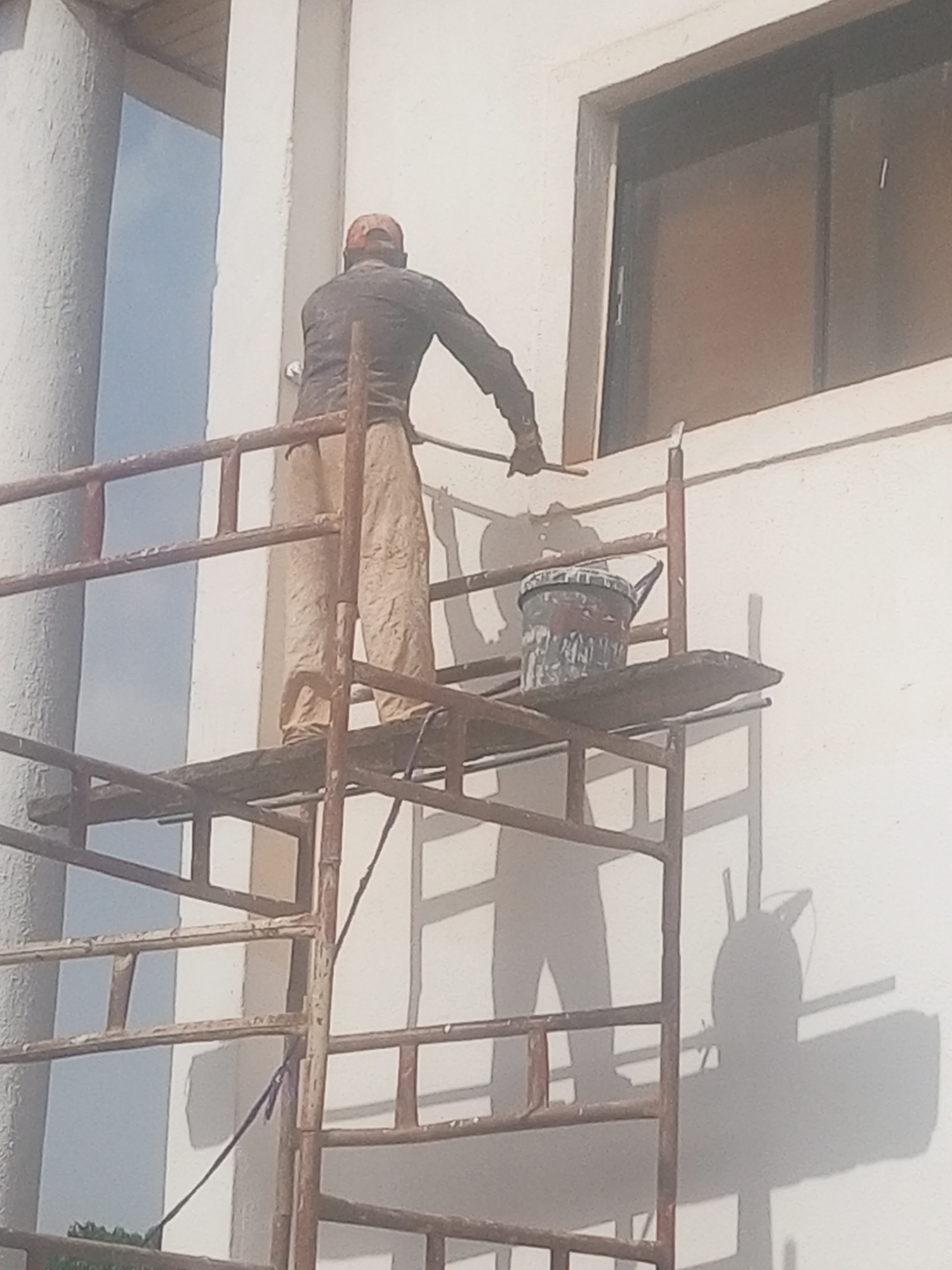 A man wearing tan trousers, a long-sleeved shirt, and a cap stands on a rusty metal scaffolding in Yelwa, Nigeria, painting a white building around a window. A paint bucket sits on the scaffolding platform. The shadow of the man and the scaffolding are visible on the building's exterior.