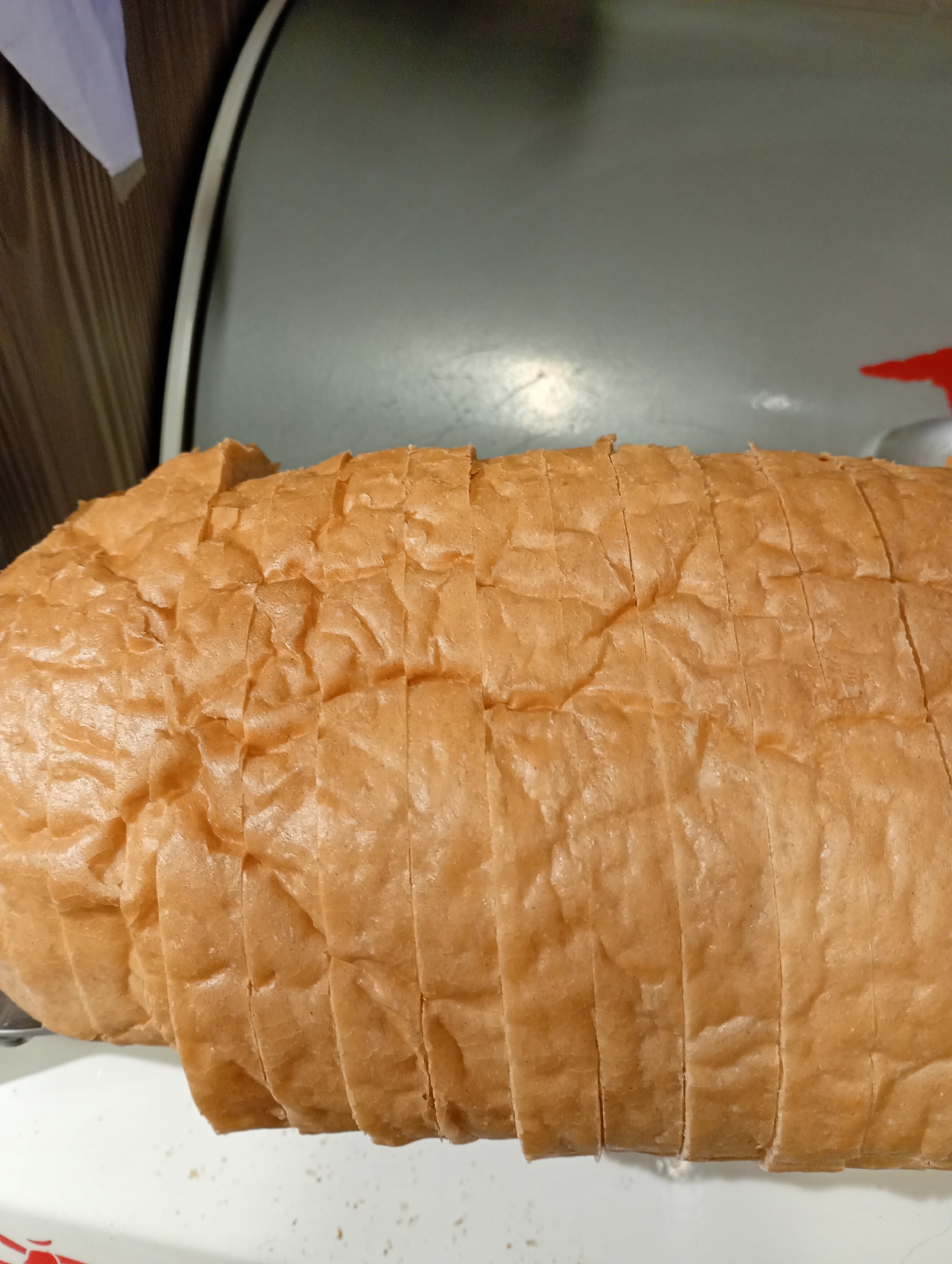 A close-up, overhead shot shows a sliced loaf of light brown bread. The bread is on a white surface, and appears to be part of a larger loaf. The crust is golden brown and slightly wrinkled. In the background, a dark gray surface is visible, possibly a countertop or appliance. A faint red object is also visible in the upper right corner of the image. The lighting is bright, highlighting the texture of the bread.