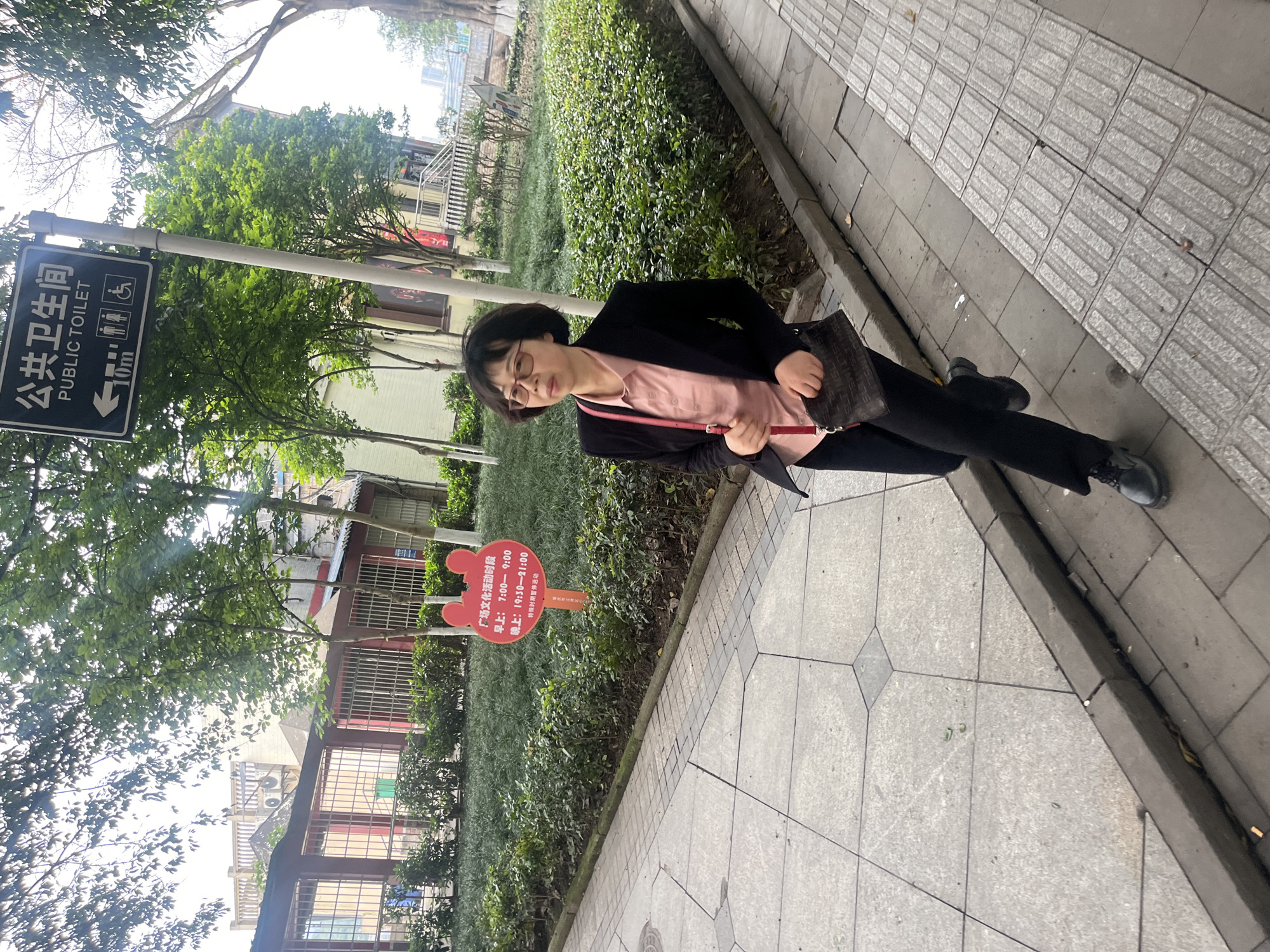 The image shows a woman standing on a sidewalk in Jiangjin, China. She is wearing glasses, a light pink shirt, a black blazer, and black pants. She is also carrying a dark-colored handbag. To her left is a sign indicating the location of a public toilet, 10 meters away. Next to her is a Mickey Mouse-shaped sign with text in Chinese. The sign indicates the hours for plaza cultural activities, with times listed as 7:00-9:00 in the morning and 19:30-21:00 in the evening. It also notes that activities are suspended during special periods. The sidewalk is made of gray concrete pavers, and there is greenery to the left of the sidewalk. The environment includes trees, bushes, and a building in the background.
