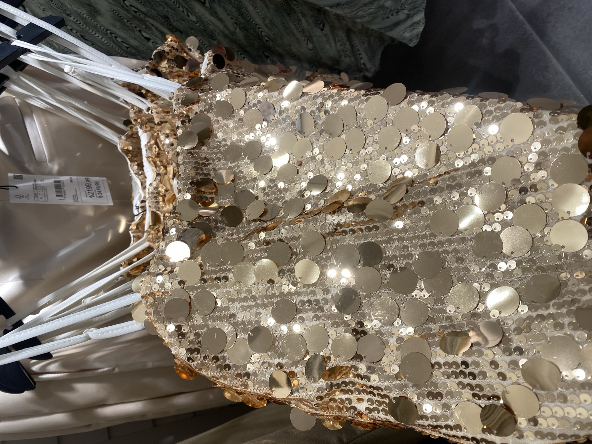 The image presents a close-up, high-angle view of a dazzling sequined garment, likely a top or dress, suspended on white clothing hangers within a retail environment. The garment is a metallic gold or champagne color, densely embellished with two sizes of round sequins: larger, flat discs and smaller, interspersed sequins, all affixed to a translucent mesh fabric. This heavy sequin embellishment creates numerous bright reflections from the lighting.
Multiple thin, white spaghetti straps are visible, gathered together by the white plastic or wire hangers. Attached near the straps is a white price tag. Clearly legible on the tag are two prices: "₺2419.99" (Turkish Lira, consistent with the Alanya, Turkey location) and "฿2199.99" (Thai Baht). A barcode and some smaller, illegible text are also present on the tag.
The background is softly blurred, showing indistinct shapes and textures of other clothing or fabrics, suggesting a typical retail store setting. No people are visible. The scene captures a festive or evening wear item on display under bright, likely artificial, lighting.