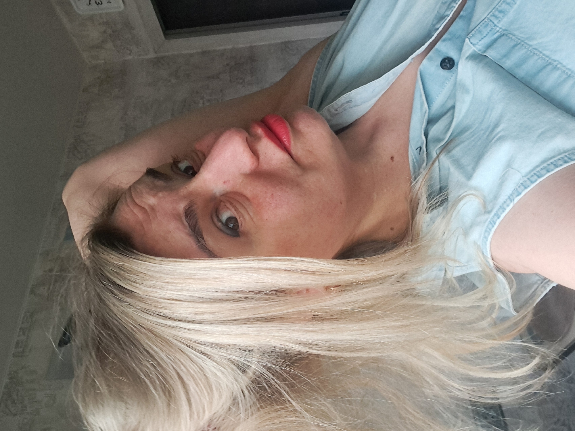 A woman with blonde hair and red lipstick is posing for a selfie indoors. She is wearing a light blue shirt. The background is a grey wall with a small white clock on the upper left corner, above what seems to be a dark-colored cabinet or window frame. There is no information in the image to indicate a location in the city of Elektrostal, Russia.