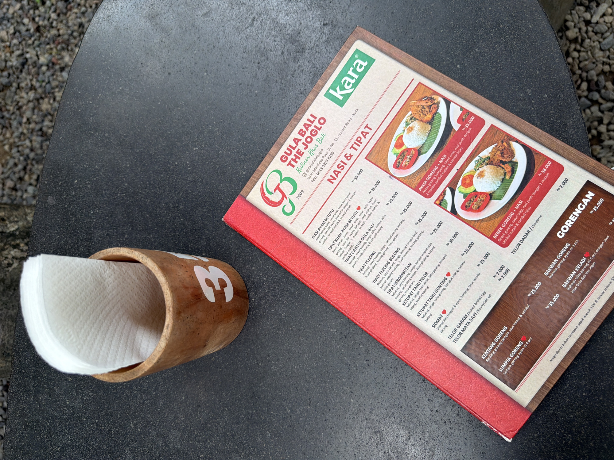 The image shows a menu for Gula Bali The Joglo, a restaurant located at Jalan Merdeka Raya VI No. 11, Sunset Road, Kuta, Bali, Indonesia. The menu is red and features various Indonesian dishes. It is placed on a dark, textured outdoor table, next to a wooden cup filled with white paper napkins. The wooden cup has the number "3" painted on it. In the background, gravel can be seen. The lighting suggests it is daytime. The menu has sections for "NASI & TIPAT", "GORENGAN", and other food items with their prices listed in Rupiah. Two plates of food, one appearing to be "AYAM GORENG + NASI" and another "BEBEK GORENG + NASI", are visible on the menu.