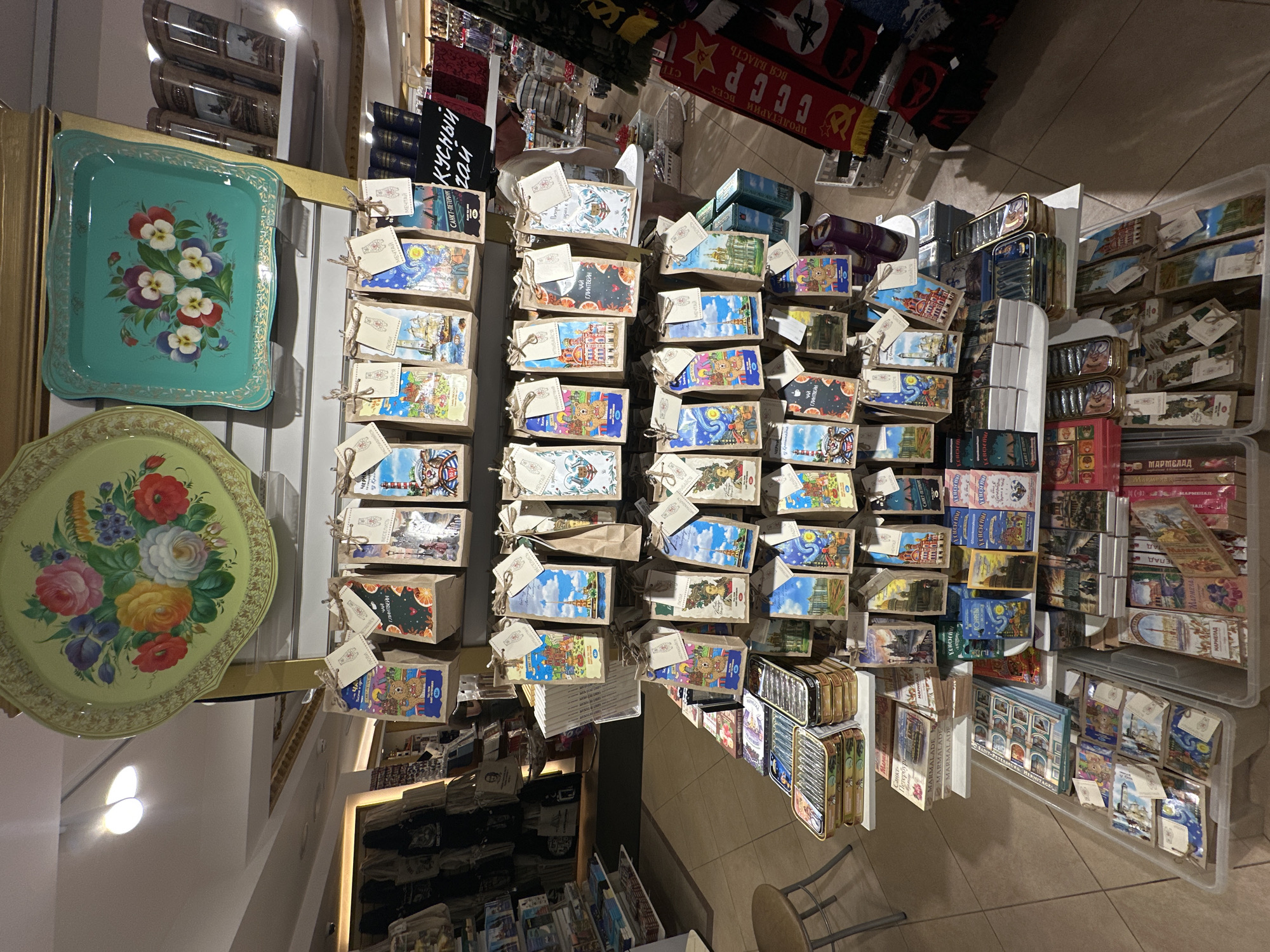 This image shows a shop in St. Petersburg, Russia, likely a souvenir shop due to the types of items displayed. From an overhead angle, the layout of the shop is apparent. There are metal trays with floral designs displayed on shelves to the left. A central table is packed with items, many of which appear to be small boxes with various designs featuring Russian landmarks or themes. Each box has a small tag attached. The boxes are neatly stacked in rows and columns. To the right, items are organized in clear plastic bins. The Russian flag with the communist symbol, other scarfs and memorabilia are seen on top shelves.