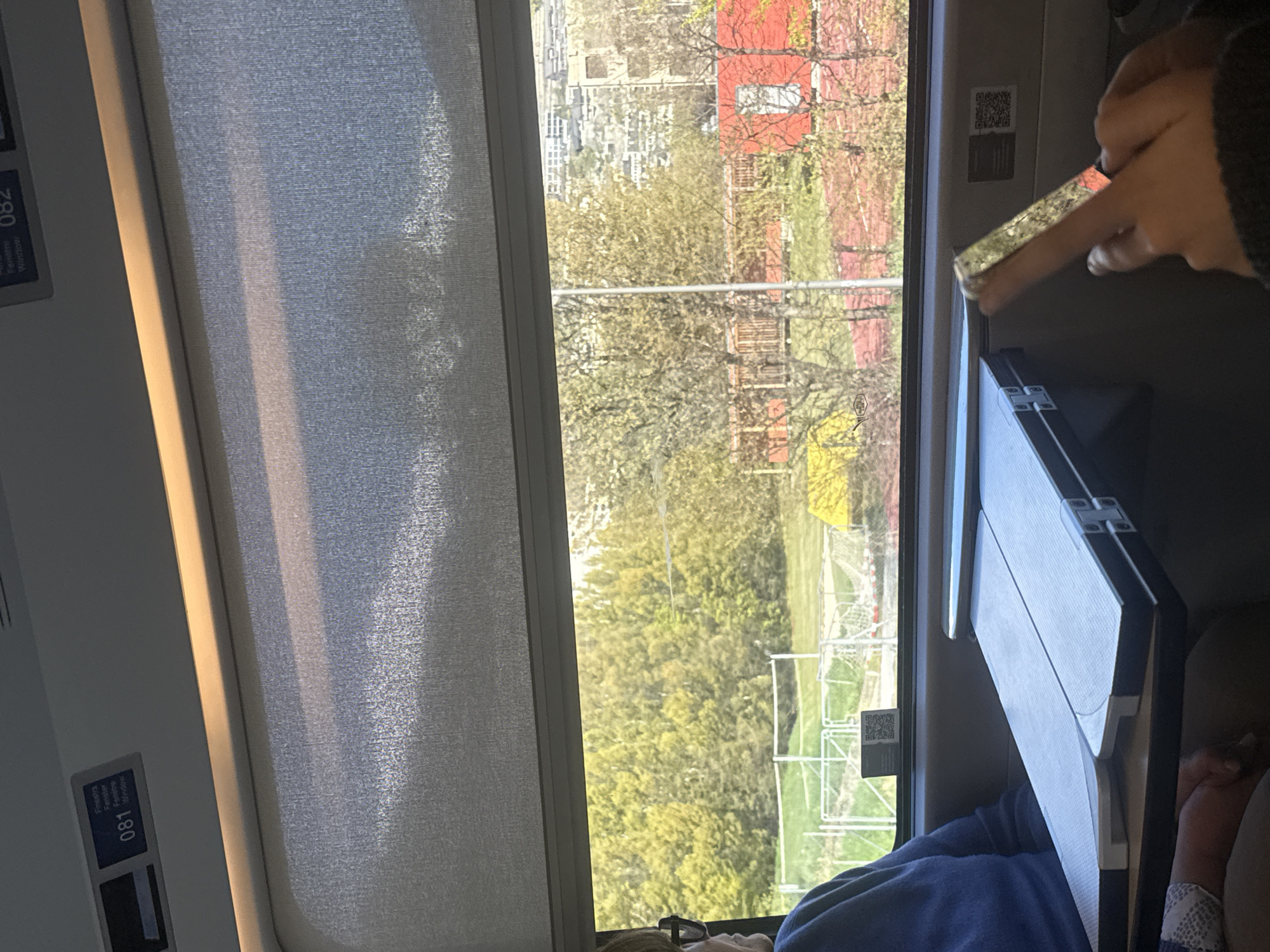A view from inside a train carriage traveling through Sion, Switzerland. A window shows an exterior view of a city with a mix of residential buildings and green areas. Trees with green and yellow foliage are visible. A partially closed blind covers the window. Inside the train, a fold-down table is open and a person wearing a blue shirt is seated by the window. The fingers of another person’s hand are visible, reaching to the window. Small signs with numbers “081” and “082” are on the wall.