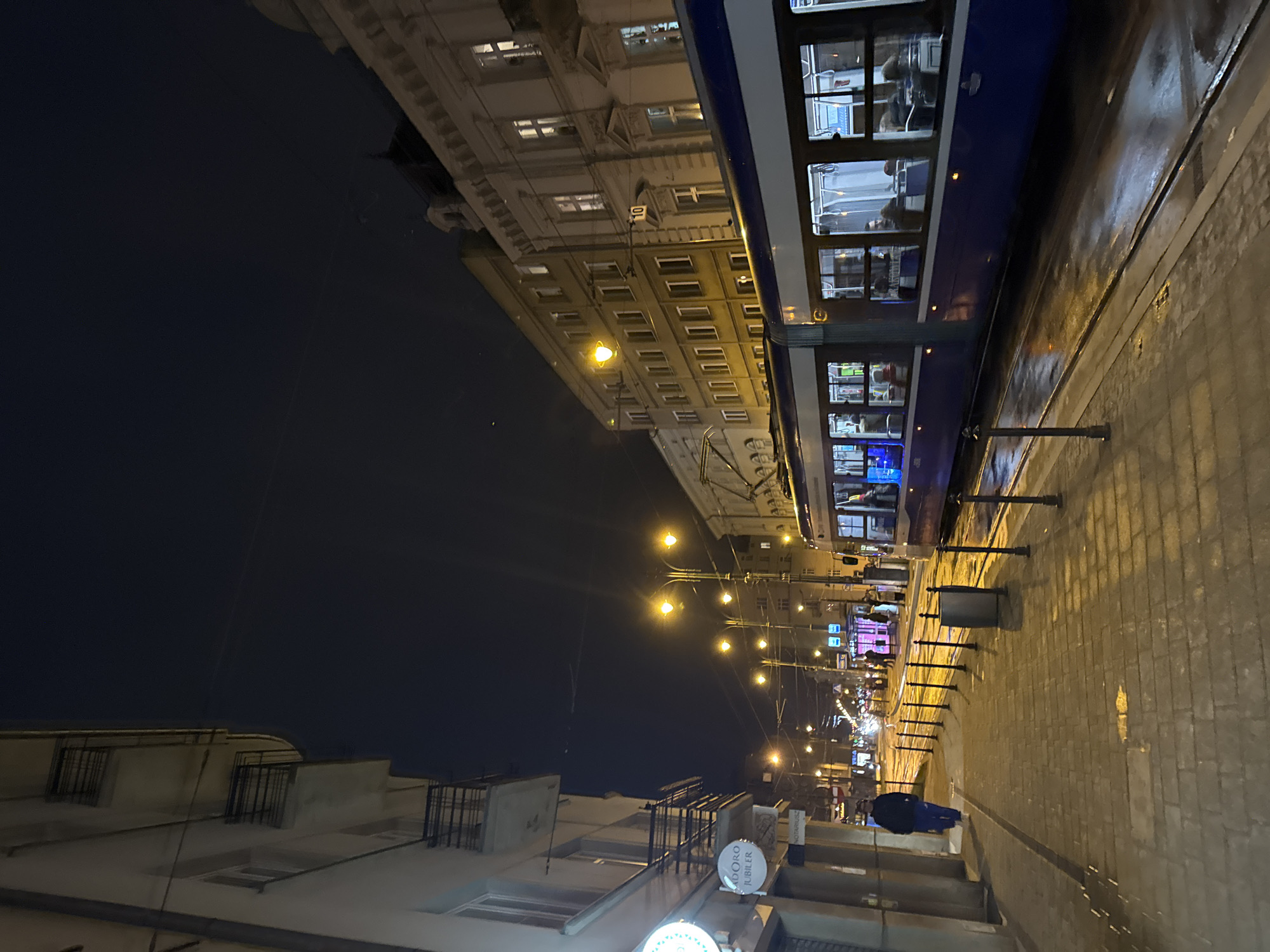 The image depicts a street scene in Krakow, Poland, at night. A blue and white tram is partially visible on the right side of the image, with passengers discernible through the windows. The tram runs parallel to a brick-paved sidewalk. Buildings line both sides of the street, with ornate facades and numerous windows illuminated by interior lights and street lamps. A series of evenly spaced lampposts cast a warm yellow glow along the street. In the foreground, a person wearing a blue jacket is present. Signage and advertisements are visible on the lower left. The sky is dark, suggesting it is late evening or night. The overall environment appears urban and well-lit.