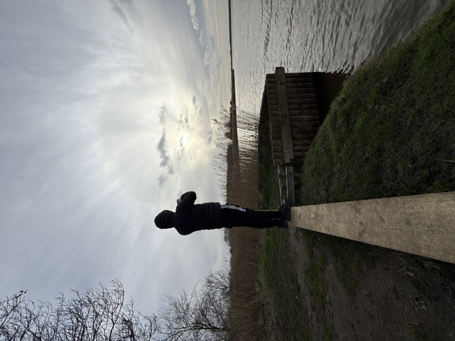 A lone individual stands balanced on a weathered wooden plank extending from a grassy embankment towards a body of water. The person, viewed from behind, wears a dark hooded puffer jacket, dark trousers with a white vertical stripe on the outer leg, and dark athletic shoes. They are holding an object, likely a mobile phone, with both hands, raised to capture an image or video. Adjacent to the plank and to the right, a wooden piling structure forms part of the bank's edge. The expansive body of water exhibits surface ripples and reflects a bright, overcast sky. The distant banks are lined with dense, tall, light-brown reeds. Bare deciduous tree branches are visible on the far left. The sky is predominantly overcast with wispy cloud formations, but the sun is visible breaking through the clouds near the horizon, creating a bright glare on the water. This environment is characteristic of the waterways and wetlands found within the Norfolk Broads, a national park area that extends into the North Norfolk district, United Kingdom.