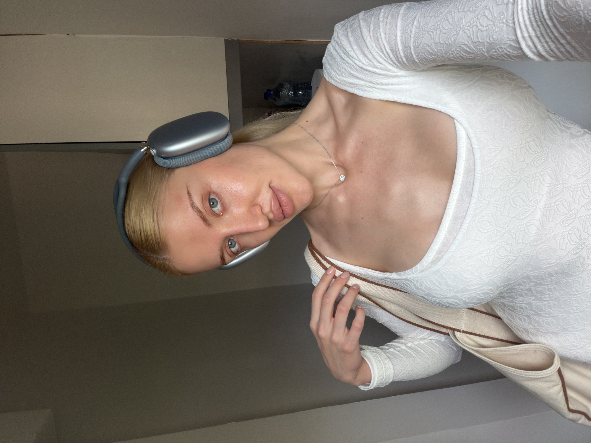 A fair-skinned woman with blonde hair is the primary subject of this shot. She is wearing gray headphones, a white long-sleeved dress, and a silver necklace with a small pendant. A cream-colored bag with brown trim is slung over her shoulder, and she is holding the strap with her right hand. The background is composed of neutral-colored walls, with an alcove above her head containing a water bottle. The location is uncertain and cannot be determined to be Belgrade, Serbia from this image.