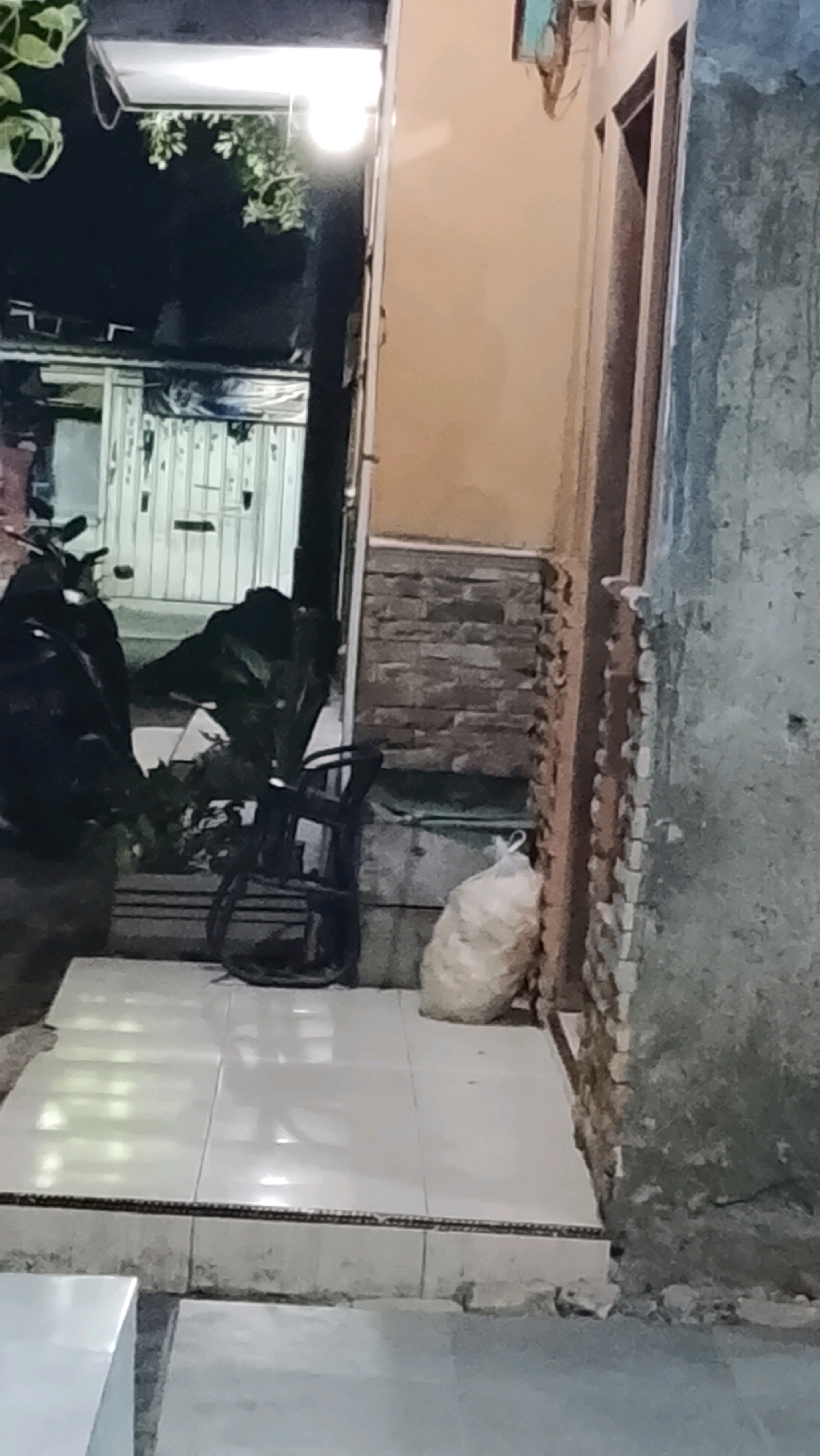 The image shows a dimly lit alleyway or narrow passage between buildings in Ujungrusi, Indonesia. On the left side, a motorcycle is parked next to a white door with vertical slats. A bright light hangs overhead, illuminating some green foliage. In the center, a black chair with a woven seat is placed on a tiled floor. To the right of the chair, a white sack of material is leaning against a wall constructed of rough, irregular stones. The wall on the far right appears to be weathered and gray. The overall impression is one of a somewhat cluttered and utilitarian outdoor space.
