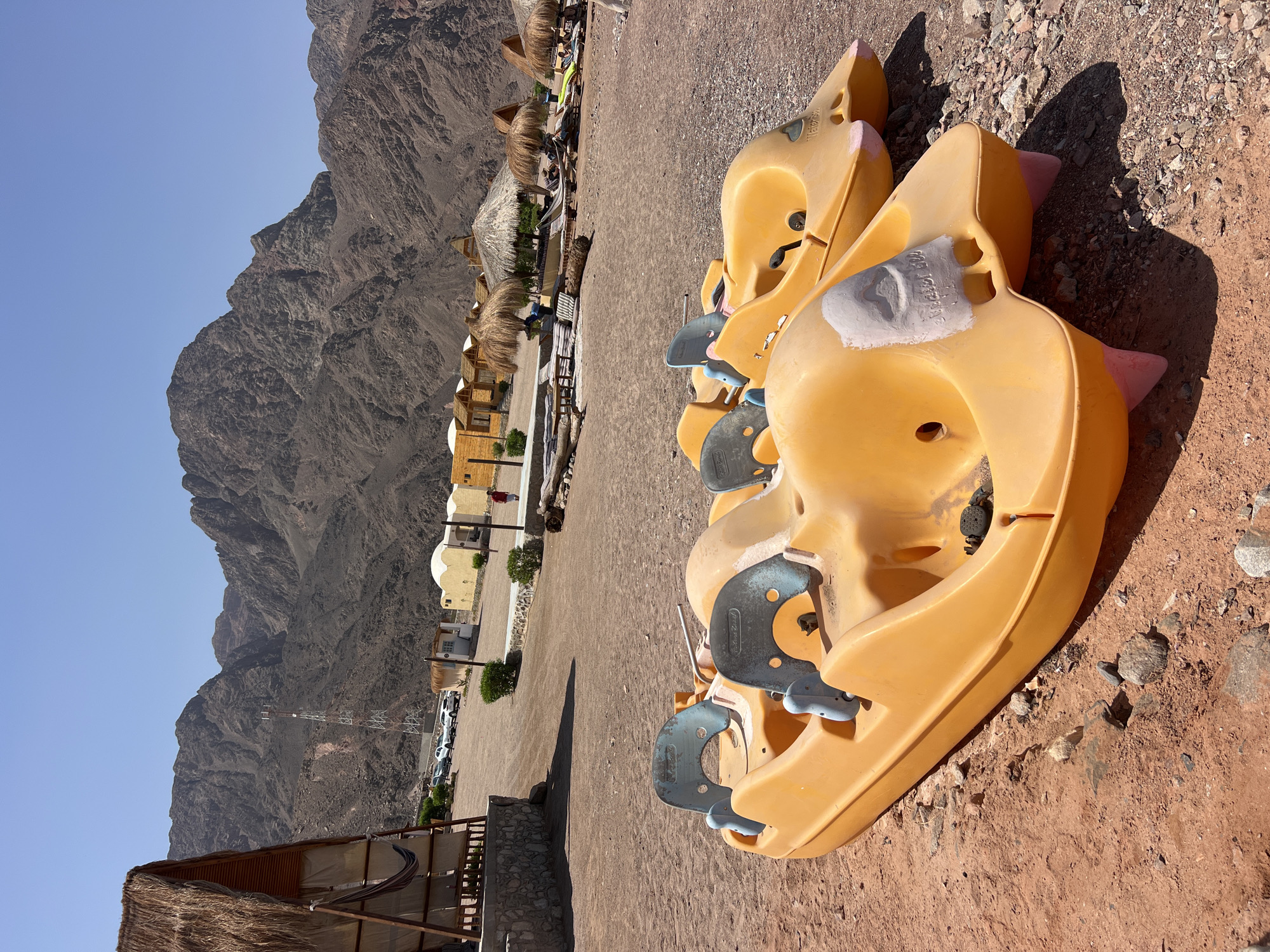 In the foreground of the image, two bright yellow pedal boats are parked on a sandy and rocky shore. The boats are side-by-side, facing towards the right of the frame. They appear to be for recreational use, with seats and steering mechanisms visible. The boat on the left has a patch of light gray material covering part of its front.
In the background, a small resort or camp area is visible. There are several small, dome-shaped structures with light-colored walls and thatched roofs, suggesting a desert or coastal location. A tall, red and white telecommunications tower stands prominently between a cluster of these buildings and the mountains. The terrain is dry and arid, with rugged, barren mountains dominating the horizon under a clear blue sky. The scene suggests a sunny day in a remote, possibly tourist-oriented location in Egypt.
