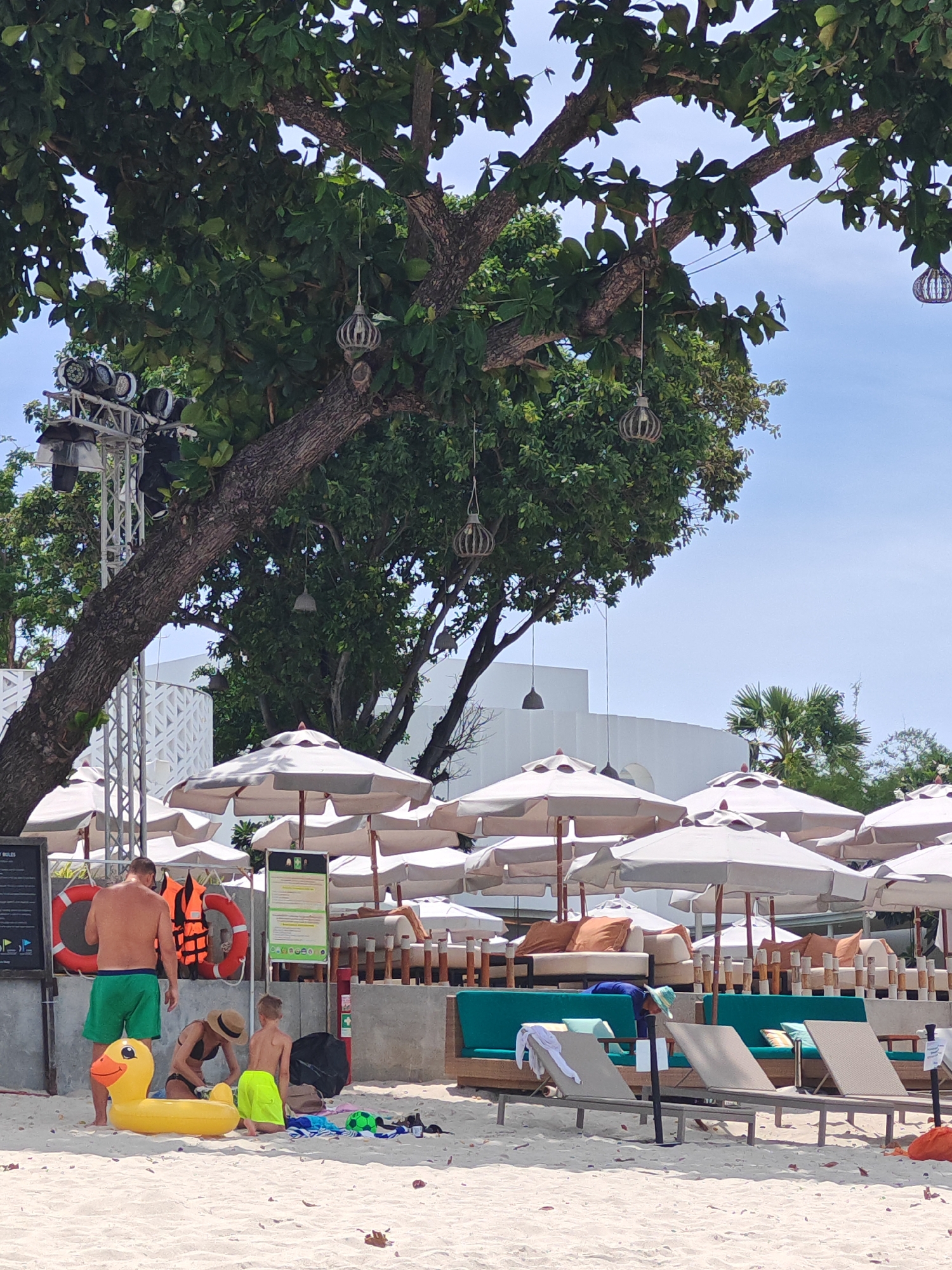 A low-angle, outdoor, medium shot captures a beach scene in Baan Chaweng, Thailand, under a clear blue sky. In the foreground, a yellow inflatable duck rests on the sandy beach. To its left, a man in green shorts stands, and next to him, a woman in a dark swimsuit wearing a straw hat kneels. A child in bright yellow-green shorts is also kneeling nearby, facing away from the camera. Behind them, a wall with information boards is visible, along with orange life preservers hanging on it. A collection of white beach umbrellas shades lounge areas with tan cushions and teal seating. Palm trees and other green foliage are visible in the midground and background, with white buildings situated further back. A large tree with exposed branches and hanging decorative lanterns extends from the upper left corner of the frame, partially obscuring the sky and the structures behind it. The scene is brightly lit by natural sunlight.