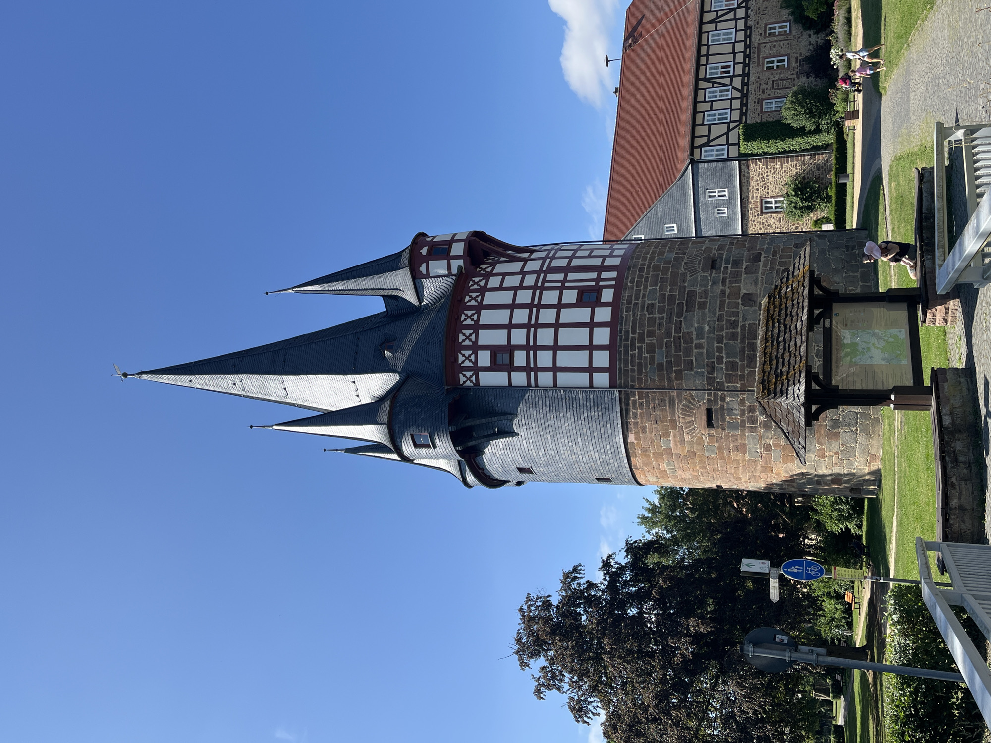 The image depicts a tall, historic tower with multiple spires against a clear blue sky. The tower has a stone base and a timber-framed upper section with white infill and dark timbers. To the right of the tower, a red-tiled roof building with similar timber framing is visible. In the foreground, a paved pathway winds through a grassy area with trees and bushes. A wooden information stand with a map is positioned near the pathway. Several people are present in the scene: a woman with a child sitting on a bench, and two women walking on the pathway in the background. The overall environment suggests a park or historical site. This is the Weiler Tor in Neustadt, Germany.