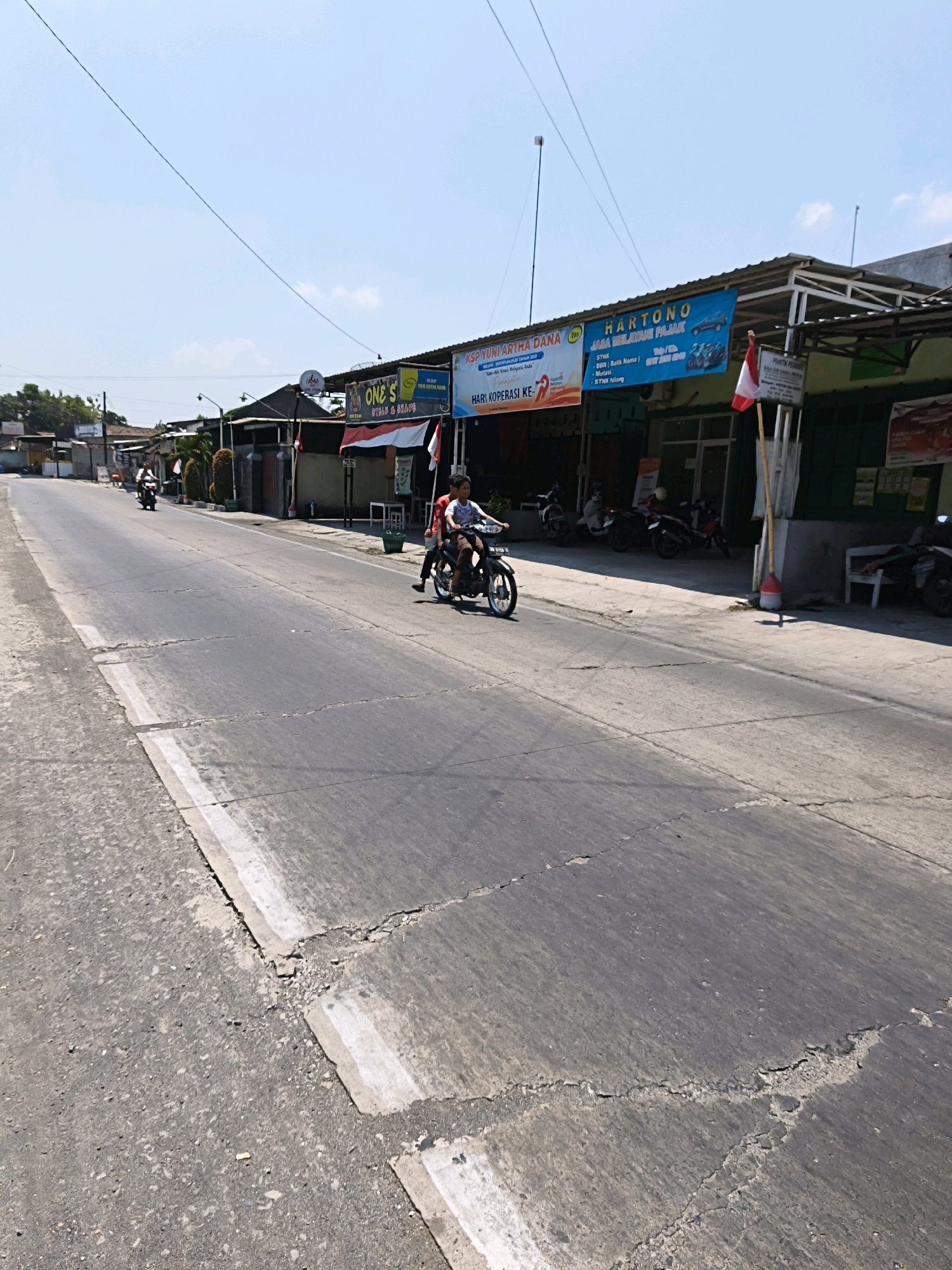 The image shows a sunny daytime scene on a paved road in Desa Dibal, Indonesia. Two people are riding on a single motorcycle, with one person driving and another person riding as a passenger behind them. The motorcycle is moving away from the viewer on the right side of the road. On the right side of the road, there are several shops with signs, including one that reads "HARTONO JASA MELAYANI PAJAK" which translates to "HARTONO TAX SERVICES". A small Indonesian flag is planted near the front of the shops. A person appears to be resting on a white bench in front of one of the establishments. There are also several other motorcycles parked in front of the shops. The road is a two-lane asphalt road with white markings. The environment is urban, with buildings lining the street and a clear blue sky overhead with some power lines visible.