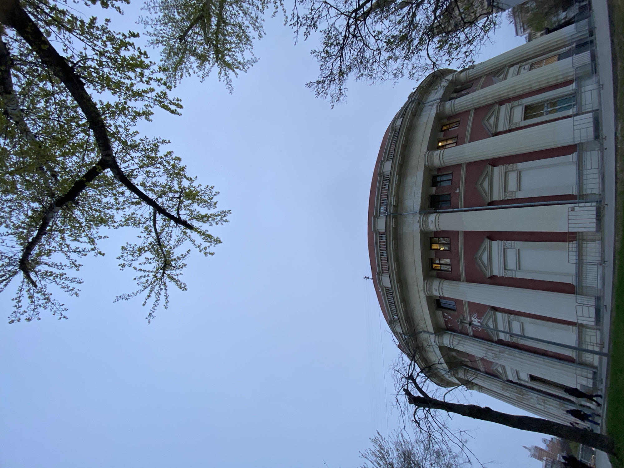 A low-angle, upward-looking photograph captures a grand, curved building framed by tree branches against a light blue twilight sky. The building, positioned prominently on the right side of the frame, features a reddish-pink facade adorned with numerous tall, white classical columns and pilasters, detailed with capitals and bases. Multiple windows are set into the facade, with some emitting warm light, suggesting it is either dawn or dusk.
The building's impressive curved architecture dominates the scene. Above and to the left, tree branches with light green, budding leaves are visible, indicating spring. Some branches, particularly in the lower left, are still bare. The sky is clear, light blue, and uniformly lit, characteristic of an overcast day or twilight conditions. A narrow strip of ground or sidewalk runs alongside the base of the building, where faint, indistinct figures of people can be seen walking.
The architectural style, with its imposing scale, classical elements, and distinctive curved shape, is characteristic of structures found in Moscow, Russia. The overall mood conveyed is one of grandeur and serenity. No visible text is present in the image.