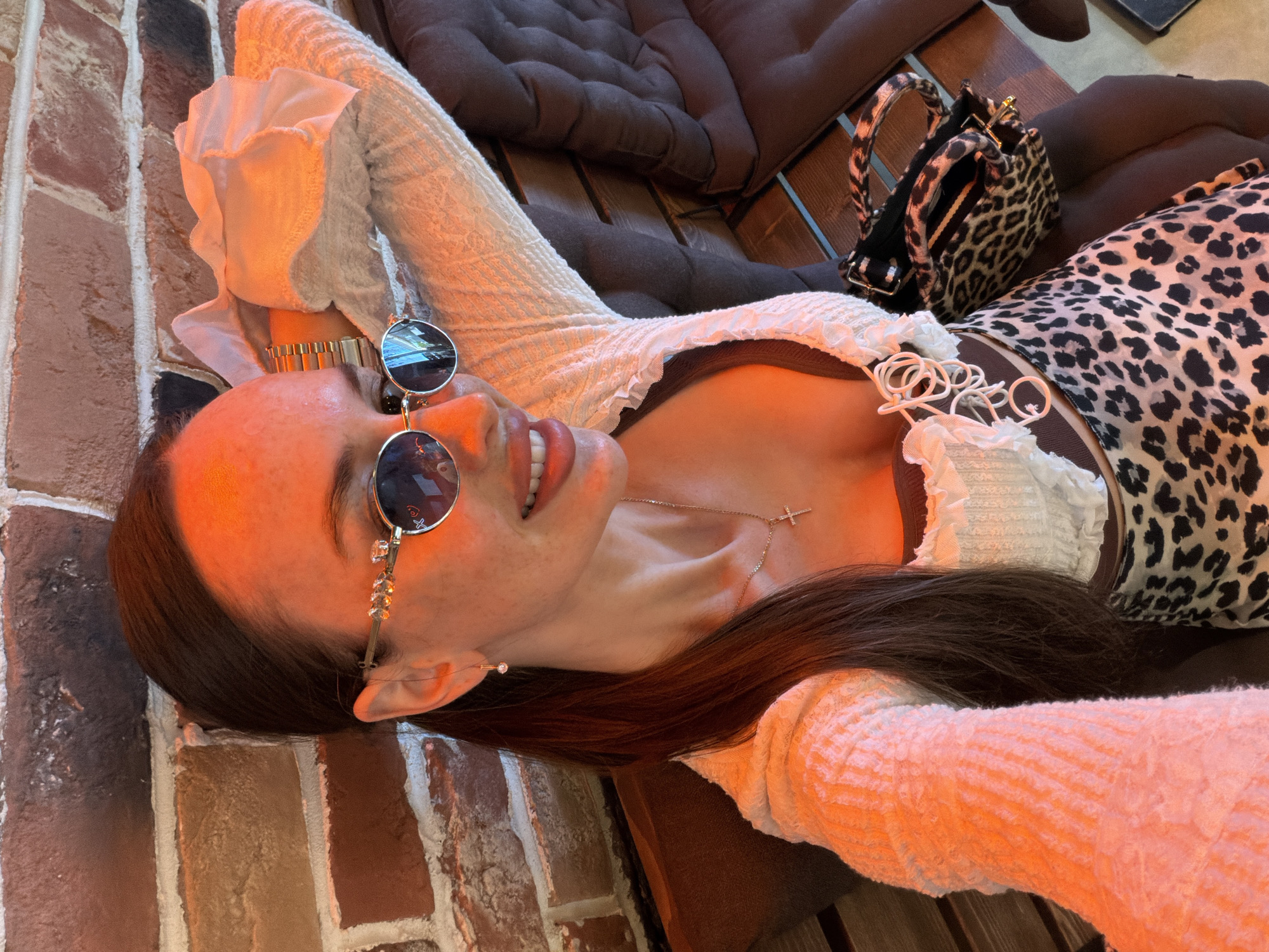 A young adult female is depicted in a selfie, taken indoors in Saint Petersburg, Russia. She has long dark hair styled in a low ponytail and visible freckles across her face. She wears round, reflective blue-tinted sunglasses with gold-toned frames embellished with clear gemstones on the temples. Her attire consists of an open, long-sleeved, textured white cardigan or shrug worn over a white top with a ruffled neckline and decorative white loops down the front, beneath which a dark brown camisole is visible. A leopard print skirt or dress with black spots on a beige background is worn on her lower body.
She is smiling toward the camera. A gold-toned chain with a small cross pendant is around her neck, and a small, clear gemstone drop earring is visible in her right ear. A gold-toned metal bracelet watch is on her left wrist. Her left arm is bent and raised, resting on a dark brown vertically tufted cushioned bench or booth seating. A folded white fabric item is draped near her left arm. A small leopard print handbag with two top handles rests on the bench beside her. The background features a reddish-brown brick wall with white mortar joints. The entire scene is illuminated by a strong, warm, orange-hued light, casting a reddish glow on her skin and the surrounding environment.