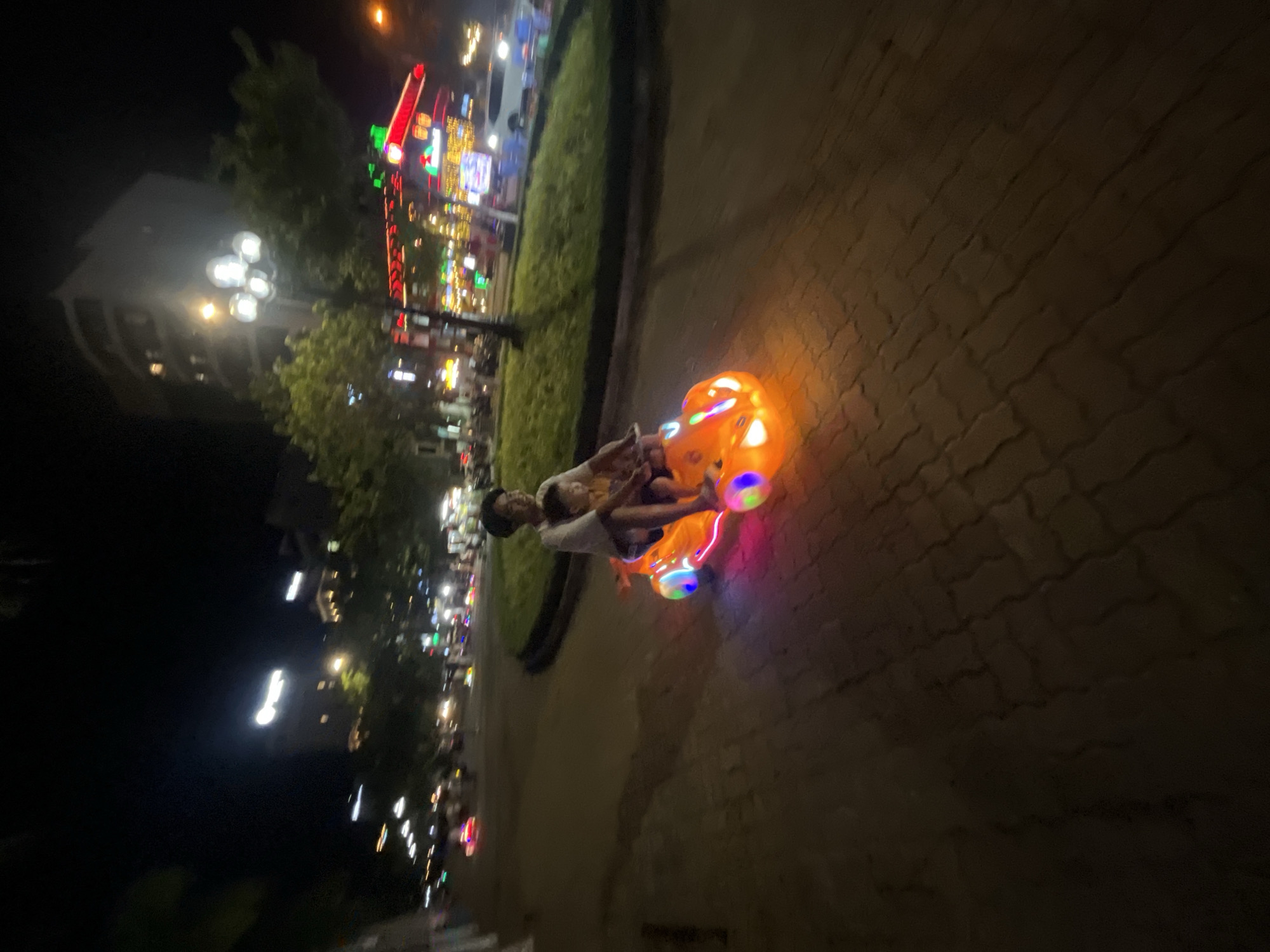 In Đà Nẵng, Vietnam, a father and his young child are enjoying a ride on a brightly lit, orange toy car at night. The car, adorned with colorful flashing lights along its sides and front, is positioned on a brick-paved pathway. The father, wearing a white t-shirt and dark shorts, is seated in the back, while the child, also in light-colored clothing, sits in front of him. They are smiling and appear to be having a pleasant time together. The background reveals a city street scene at night, with illuminated buildings, streetlights, and trees, suggesting they are in a public area with some traffic and activity. The overall atmosphere is one of leisure and family enjoyment amidst a lively urban nighttime setting.
