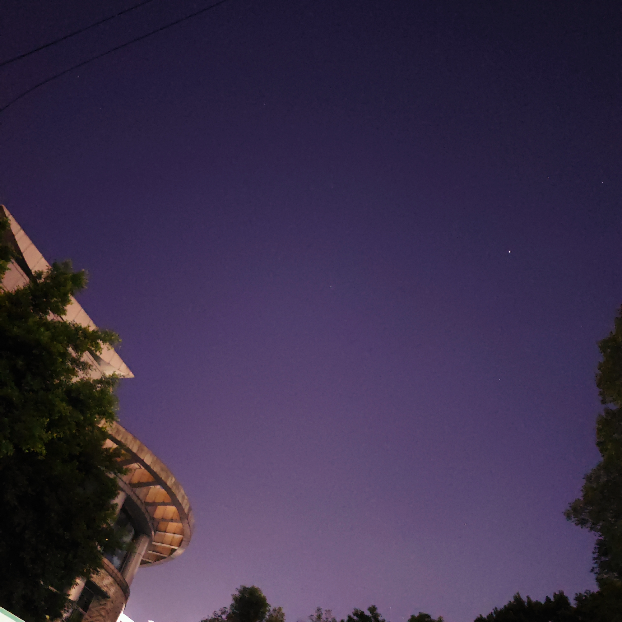 The image, taken at night, depicts a purple sky with several visible stars. The scene includes the top of a building with distinctive circular architecture surrounded by foliage, including trees. Power lines are visible in the top left corner of the frame. While the Yuzhong District, China, is provided as context, there are no specific landmarks visible to confirm a location in this district. There are no people or actions visible in the image. The scene is static.