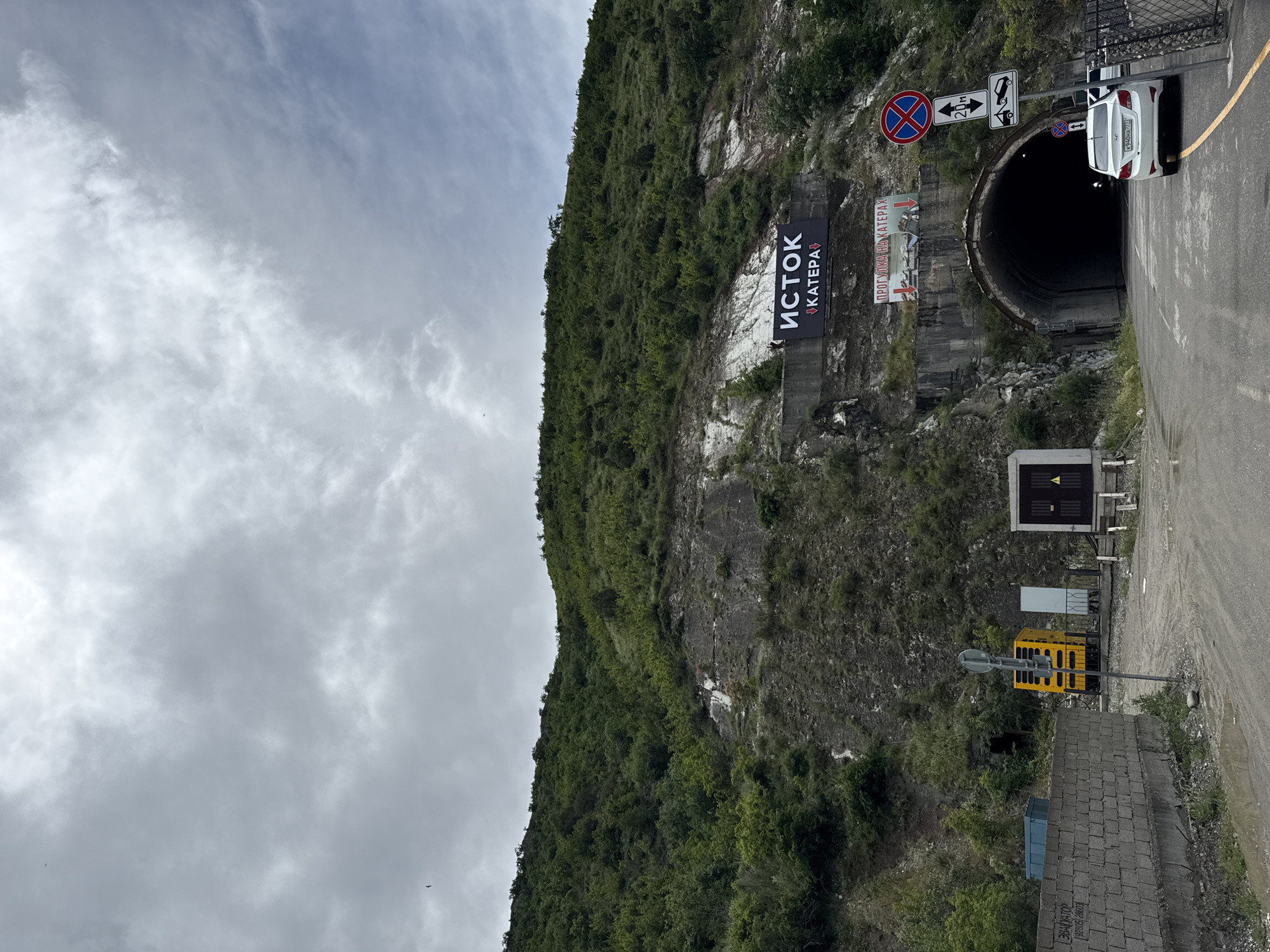 The image displays a mountainous landscape featuring a road and a tunnel entrance under an overcast sky. A large, verdant mountain covered in dense green foliage dominates the left side of the frame, with rocky sections interspersed among the greenery. The sky above is entirely covered with light grey clouds, suggesting an overcast day.
On the right side of the image, an asphalt road leads directly into a dark, circular tunnel entrance carved into the base of the mountain. A white car is partially visible, positioned near the tunnel's opening, suggesting it is either about to enter or has just exited.
Several road signs are clustered near the tunnel entrance. These include a red circular "No Stopping/Parking" sign, a white rectangular sign with an arrow and "20m," and another white rectangular sign depicting two cars, likely indicating traffic regulations. A small blue square sign with a downward arrow is also visible.
Prominently featured on the mountain face above the tunnel are two banners. A large black banner displays text in Cyrillic, reading "ИСТОК" (ISTOK) in large white letters, with "КАТЕРА" (KATERA) written below it in smaller text. Below this, a smaller banner reads "ПРОГУЛКИ НА КАТЕРАХ" (BOAT TRIPS).
Adjacent to the road on the right, there is a yellow, box-like structure, possibly equipment or a small booth, along with some concrete barriers or retaining walls. The scene depicts a functional road infrastructure integrated into a natural, rugged environment, located in what appears to be Novo-Zubutli, Russia.
