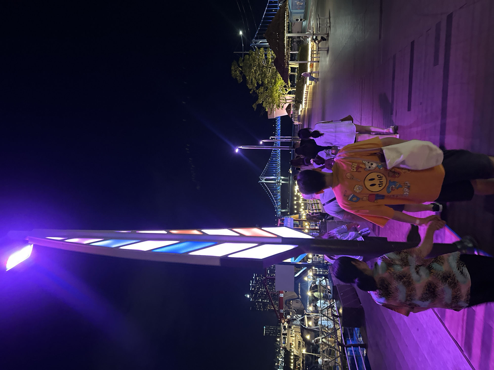 The image depicts a nighttime scene along the waterfront in Busan, South Korea, likely at The Bay 101 or a similar area. A group of people are walking along a paved promenade illuminated by vibrant purple and pink lighting emanating from a tall, decorative lamppost and the ground. In the background, a large suspension bridge, possibly the Gwangan Bridge, is visible, lit with blue lights. The cityscape with buildings and lights is also visible across the water. Several people are walking in various directions, with some walking together in pairs or small groups. There are also some seating areas with thatched umbrellas and tables. The overall atmosphere is lively and illuminated by artificial lighting.