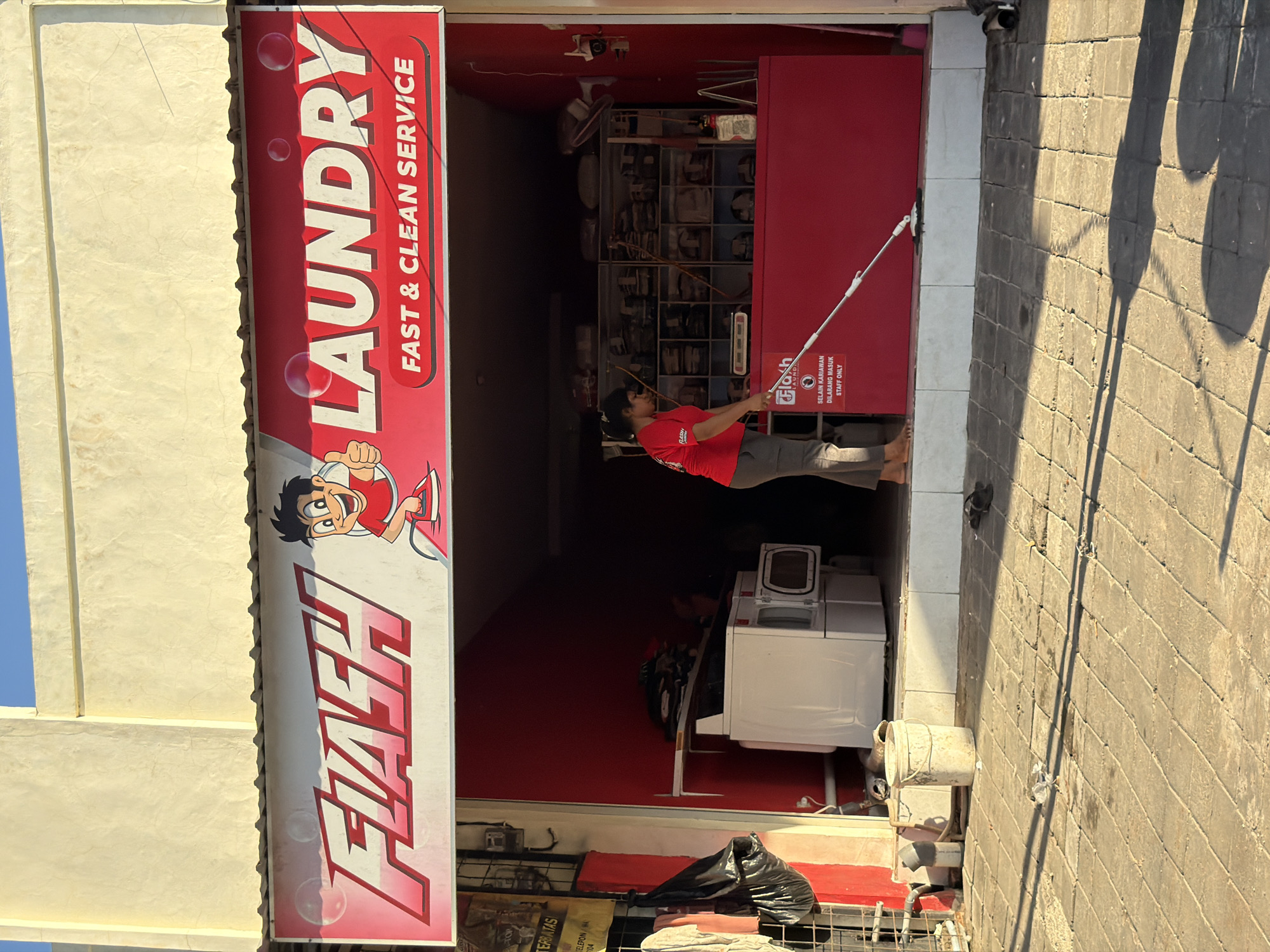 This image depicts the entrance to a laundry service business called "Flash Laundry". The storefront is painted red and features a prominent vertical sign with "LAUNDRY" in large white letters and "FAST & CLEAN SERVICE" below it. A cartoon character, also with "FLASH" in large letters, is present on the left side of the sign.
Inside the shop, a woman wearing a red t-shirt is seen cleaning the floor with a mop. Various laundry machines, including what appears to be a washing machine, are visible in the background. A sign on the red wall reads "SELAIN KARIAWAN DILARANG MASUK STAFF ONLY", indicating that only employees are allowed beyond a certain point.
The exterior of the shop is tiled, and shadows from the sun suggest it is daytime. The overall scene conveys a sense of business activity and the operation of a local laundry service.