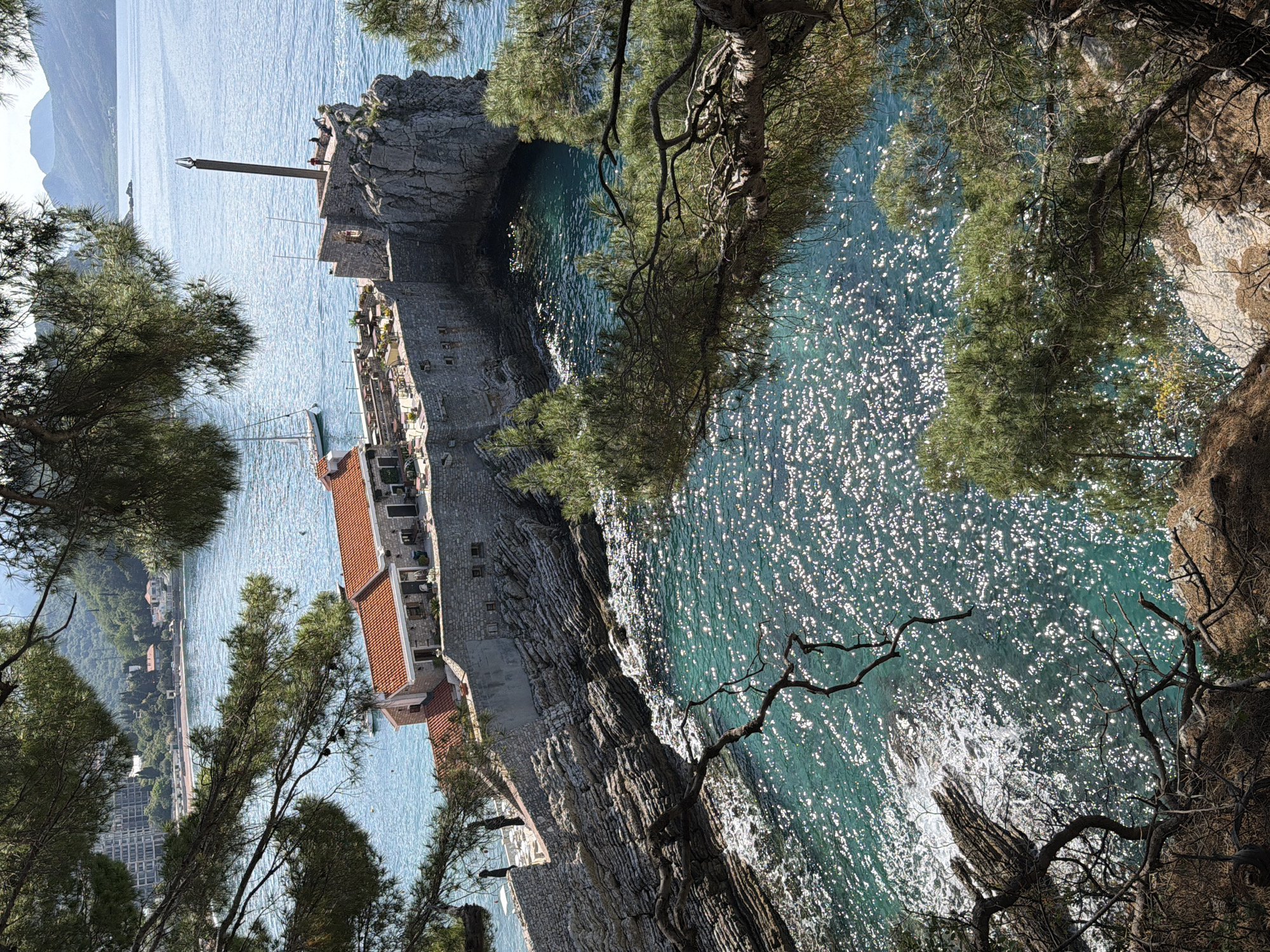 The image shows a coastal scene in Petrovac centar, Montenegro. In the foreground, trees overhang a rocky cliff. Below, the turquoise water of the Adriatic Sea sparkles with reflected sunlight. On the left side of the image, a section of the coastline features buildings with red tile roofs, constructed from stone. A tower-like structure with a pole extending upward stands at the edge of a rocky promontory. Two figures are visible atop this promontory. In the distance across the water, a town with buildings along the shoreline is visible. The sky is clear and light blue.
