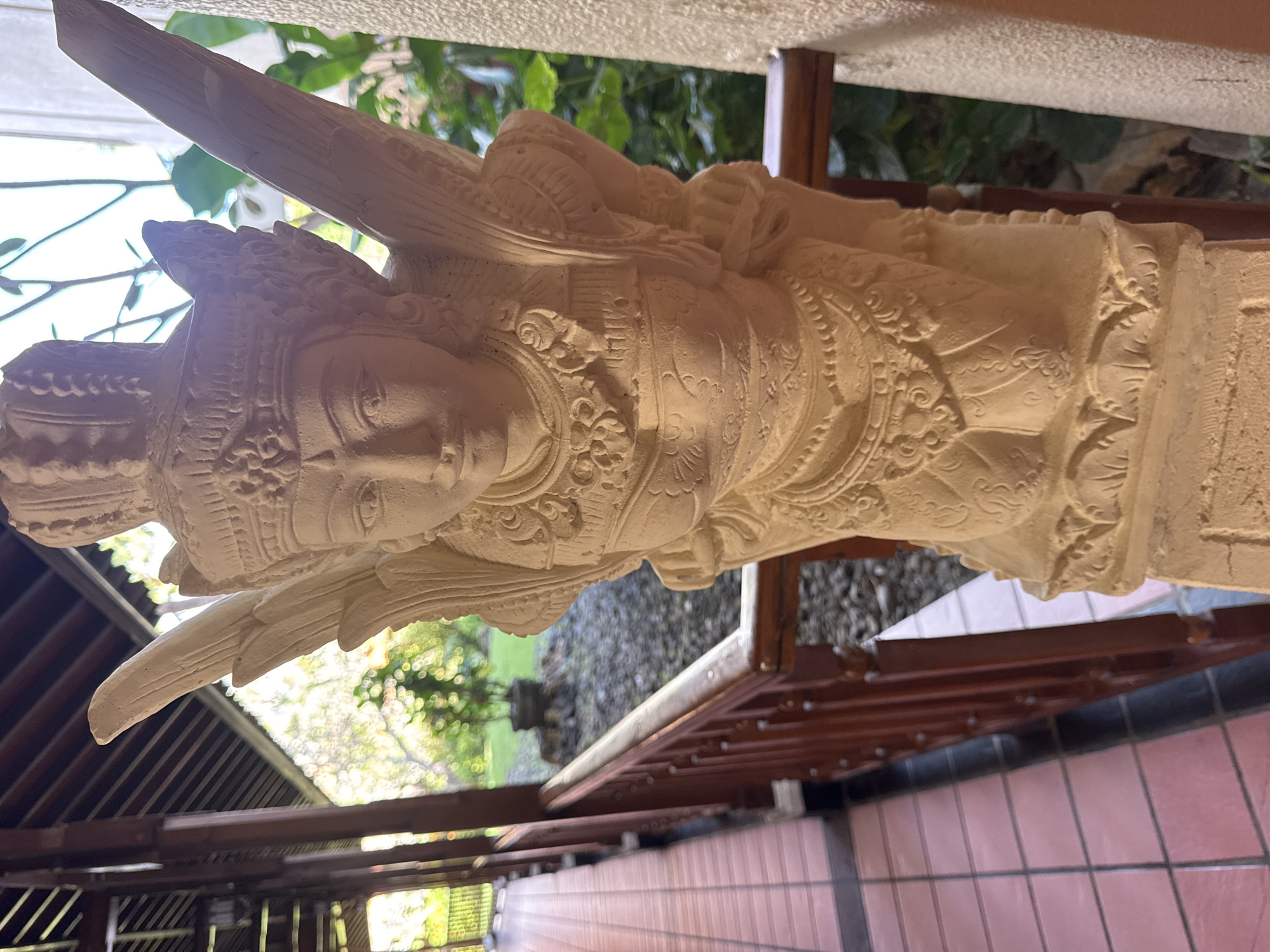 The image shows a stone statue of a winged figure in Kuta Selatan, Indonesia. The statue is light-colored and intricately carved with details on its crown, torso, and wings. It appears to be positioned on a pedestal. In the background, there is a covered walkway with a tiled floor and wooden railings. Beyond the walkway, lush green foliage and trees are visible, suggesting an outdoor setting. The lighting suggests it is daytime, possibly late afternoon, given the warm tones and shadows. The overall impression is of a cultural or decorative element within a resort or garden setting in Kuta Selatan.