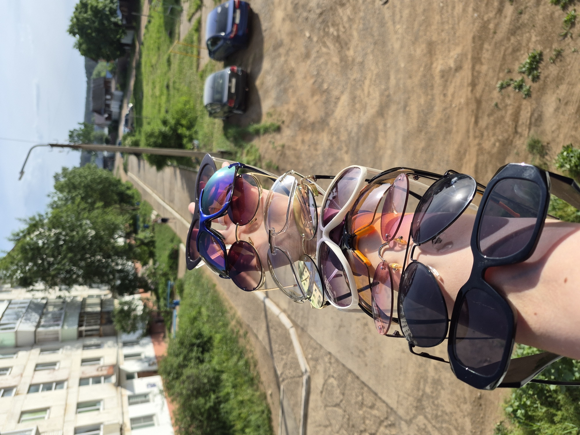 A hand holds several pairs of sunglasses, displaying them against a background of a city street in Unknown, Unknown. The perspective is slightly elevated, suggesting the photo was taken from a balcony or a higher point. A narrow street stretches into the distance, featuring two parked cars. Buildings are visible on either side of the street, interspersed with trees and greenery. The lighting indicates it is a sunny day. The sunglasses vary in style and color, arranged on a metal rack held by a fair-skinned arm.