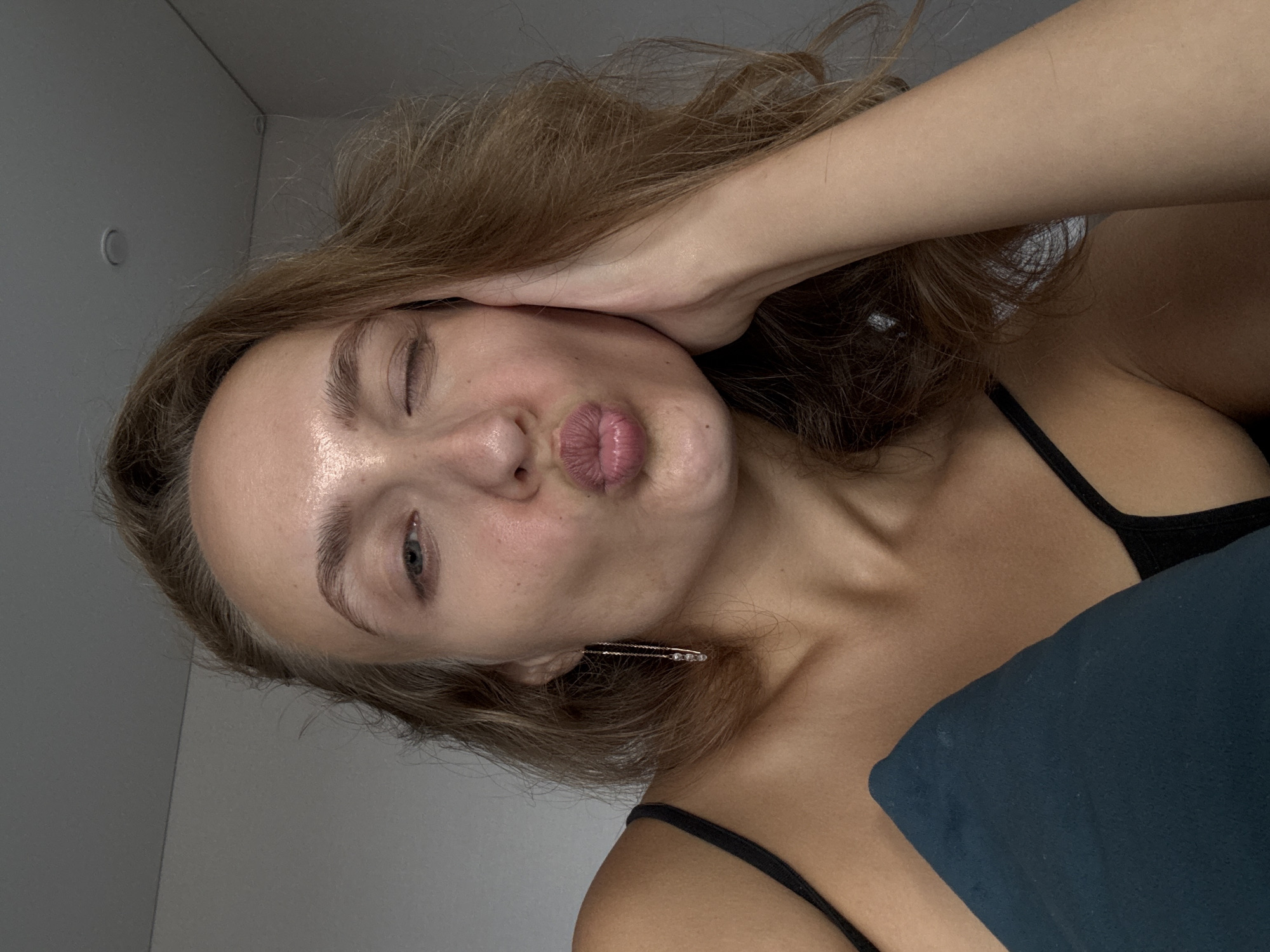 The image shows a close-up, selfie-style shot of a young woman with light brown hair. She is winking and puckering her lips for a kiss. Her left hand is resting on her cheek, and her right arm is visible in the foreground. She is wearing a black tank top. Behind her, a dark teal pillow is partially visible. The background appears to be a plain wall, suggesting an indoor setting. There is no information about the location of Подшивалово, Russia, in this image.
