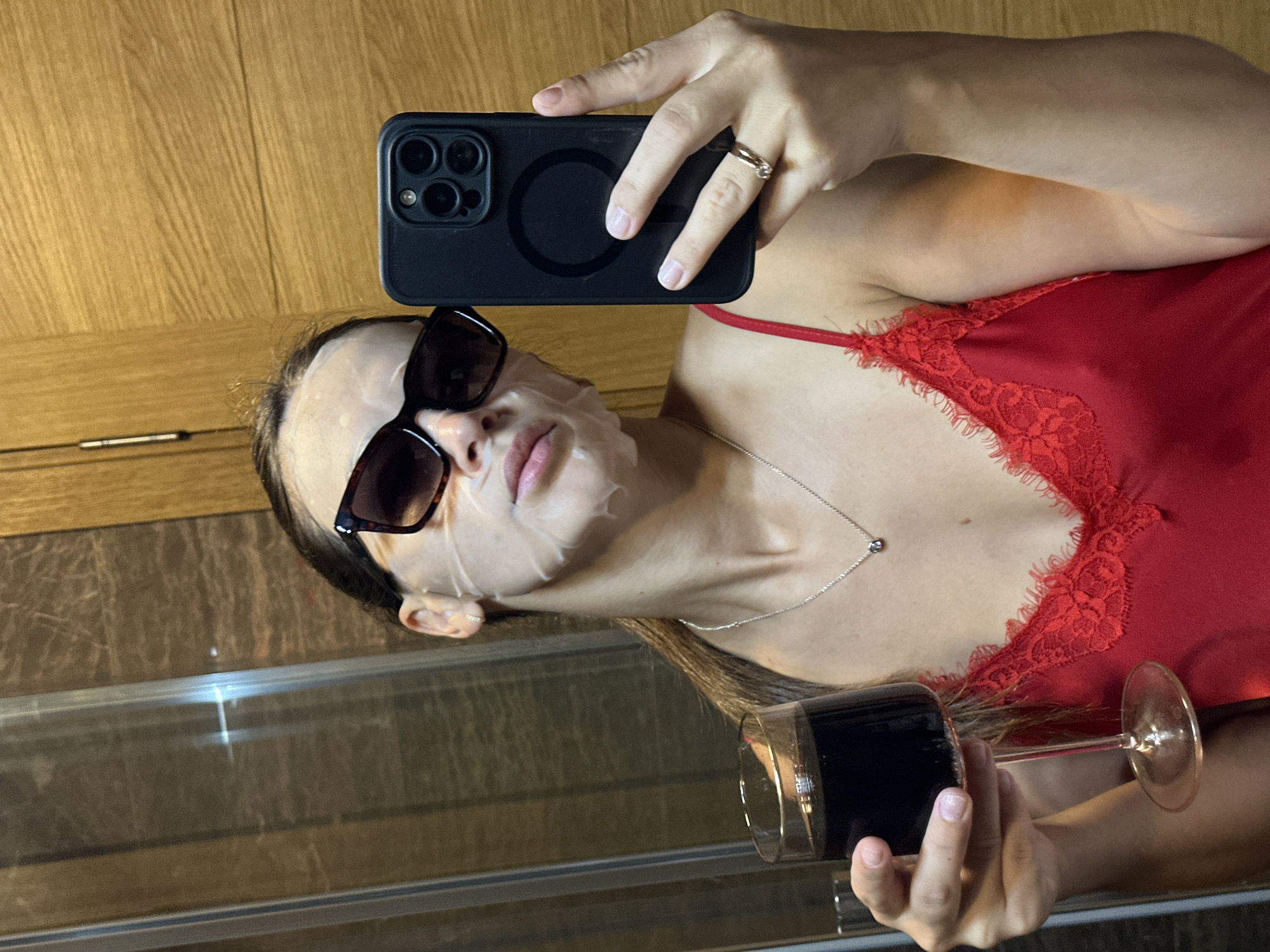 The image shows a person, seemingly a woman, holding a smartphone and a glass of dark liquid. She is wearing a red lace-trimmed top and sunglasses, with a face mask on. The scene appears to be indoors, with wooden cabinetry in the background and a mirrored surface reflecting the scene. The woman is taking a selfie with her phone, which is held up to capture her reflection. The presence of a face mask and a drink suggests a moment of relaxation or self-care. The lighting is somewhat dim, hinting at it being nighttime or an indoor setting with artificial light. The overall mood is casual and personal. There is no visible text in the image.