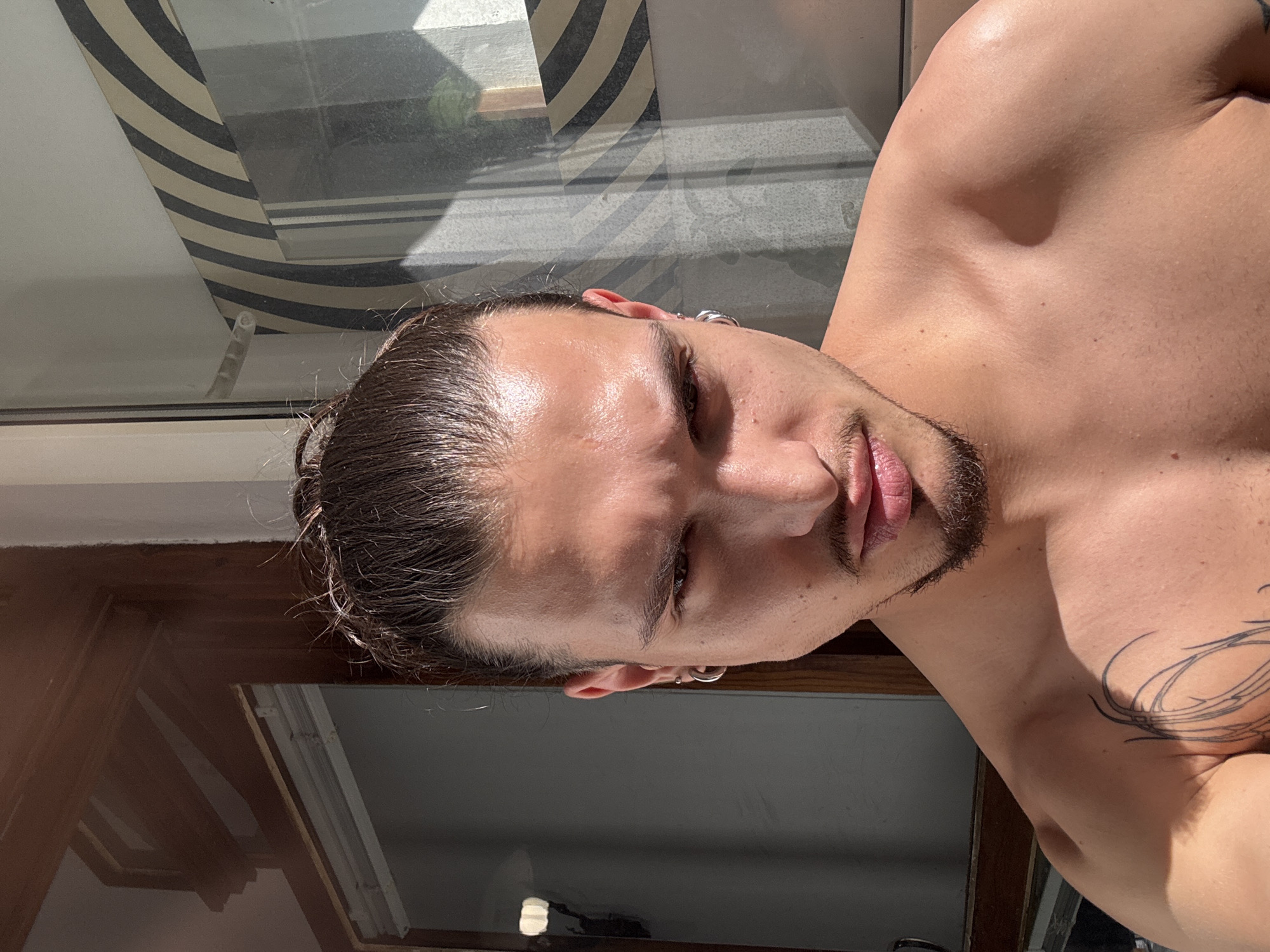 A close-up, chest-up, head-and-shoulders shot captures a young man with light skin, dark hair pulled into a bun, and a goatee. He wears hoop earrings in both ears and has a tattoo on his left shoulder and upper chest. The man is looking directly at the camera with a serious expression. The lighting suggests it is daytime, with strong sunlight casting shadows across his face and body. The background appears to be an indoor setting with large windows, one of which has a striped pattern visible. The window on the left reflects some of the interior, including what looks like blinds. The overall scene suggests an informal setting, possibly a home or studio. The man is shirtless, revealing his musculature.