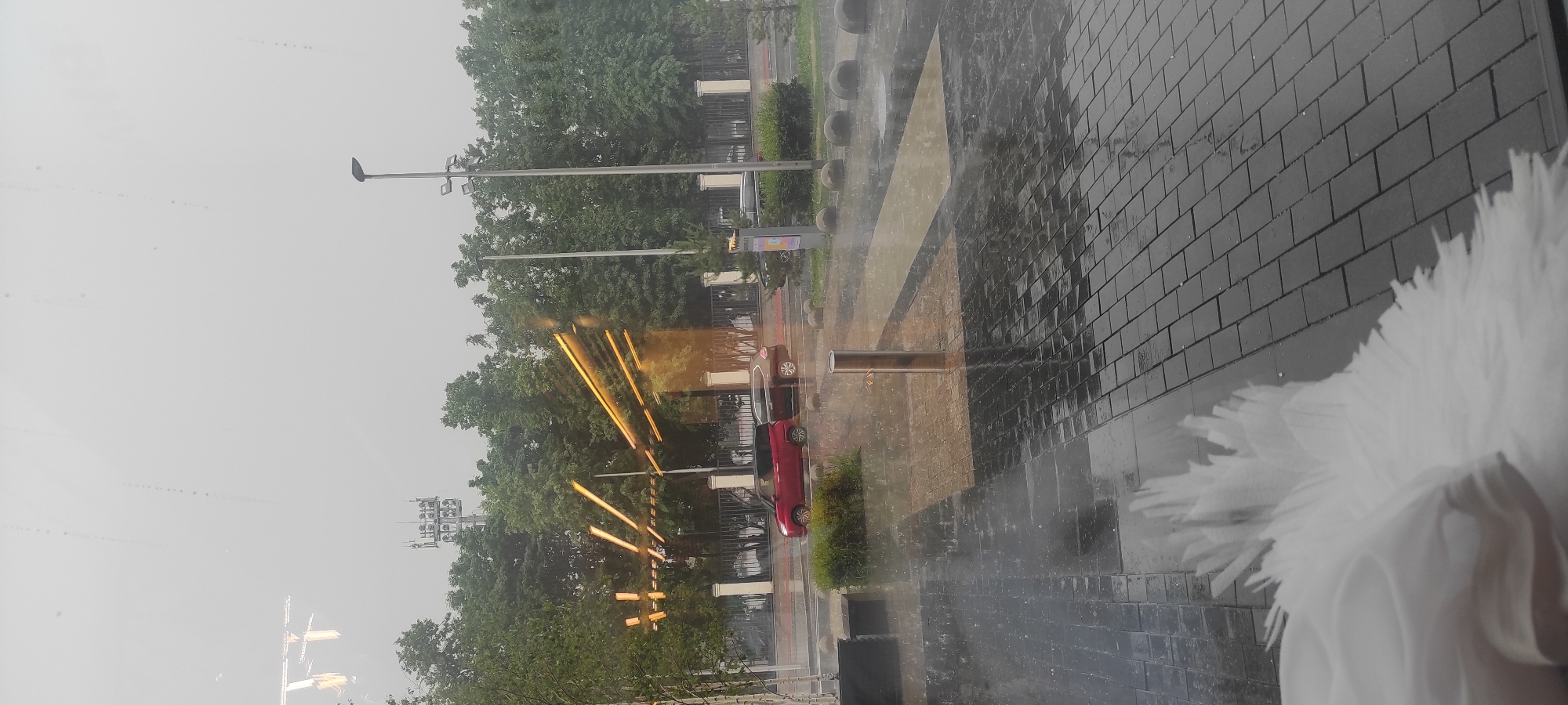 The image depicts an outdoor scene on a rainy day, likely in Moscow, Russia, given the presence of telecommunication towers and architectural styles. In the background, a tall telecommunication tower with multiple platforms and antennas is visible against a hazy sky. Several streetlights with multiple fixtures are positioned along a road.
In the middle ground, two cars are parked on the side of a road. A red car is in the foreground, and a darker colored car is behind it. A white fence runs along the edge of the property, separating it from the road and sidewalk. Trees with lush green foliage are abundant, forming a dense line behind the fence.
The foreground shows a wet, paved area with dark grey brickwork, indicating it is likely a pedestrian walkway or plaza. Puddles and reflections are visible on the wet surface, a common sight during rainfall. Several spherical bollards are placed along the edge of the paved area. A chrome-plated cylindrical object, possibly a bollard or a part of street furniture, stands in the center of the paved area, reflecting the surrounding environment.
In the bottom left corner, a blurred white object, resembling a large feather or a piece of fabric, is prominently in the foreground, possibly indicating the perspective is from inside a building looking out. The rain is clearly visible as streaks and splashes on the ground and in the air. The overall atmosphere is overcast and damp, characteristic of a rainy day in an urban setting.