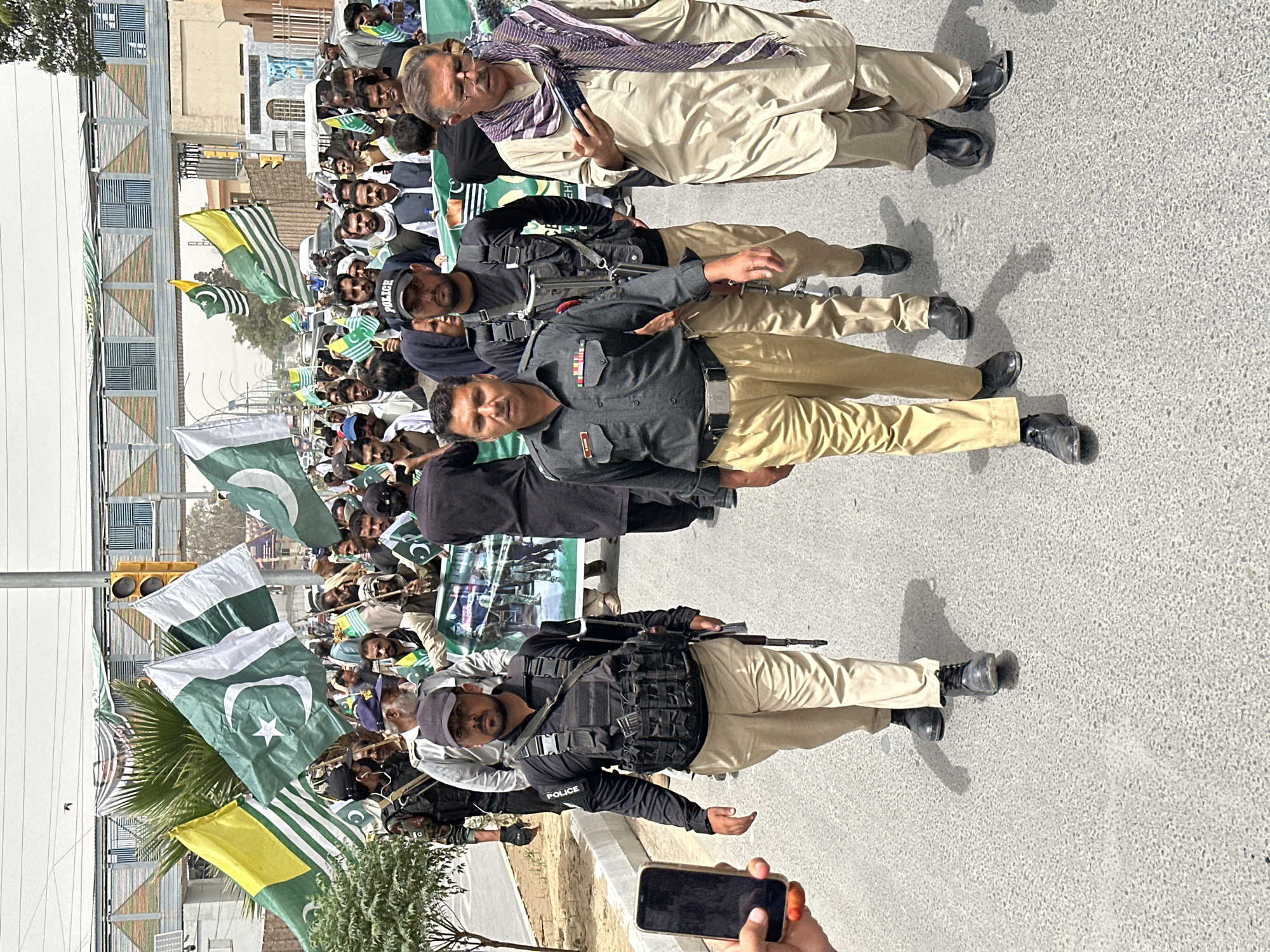 The image depicts a street scene in Quetta City Tehsil, Pakistan, where a group of people are participating in a procession. Several individuals are seen carrying flags, including the national flag of Pakistan and flags of what appear to be regional or political groups, characterized by green and white colors with a crescent and star. Some individuals are wearing police uniforms, including one in the foreground with a "POLICE" patch on his sleeve, armed with a rifle. Another person in a police uniform is to the left, wearing a tactical vest with multiple pouches. A man in civilian clothing, wearing a light-colored shalwar kameez and a scarf, is walking towards the right, looking at a mobile phone. The background shows buildings, a traffic light, and an overhead pedestrian walkway. The overall atmosphere suggests a public gathering or demonstration.