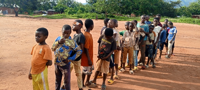 The scene depicts approximately 18-20 children, predominantly boys, standing in a loose queue on reddish-brown earth in Ngalula, Democratic Republic of the Congo. The children, appearing to range in age from roughly 5 to 12 years old, are of African descent and wear varied, casual clothing including t-shirts, patterned shirts, shorts, and long pants. Some wear sandals or flip-flops, while others are barefoot. One child has a white band tied around his forehead. Most individuals are looking towards the left or slightly ahead of the camera's perspective. The ground is unpaved, sun-baked dirt, with distinct shadows indicating bright daylight. In the background to the left, sparse trees and several simple, single-story structures with dark roofs are visible. To the right and in the far background, dense green foliage and distant hills or mountains are present under a clear sky. There are no overt interactions observed between the individuals.