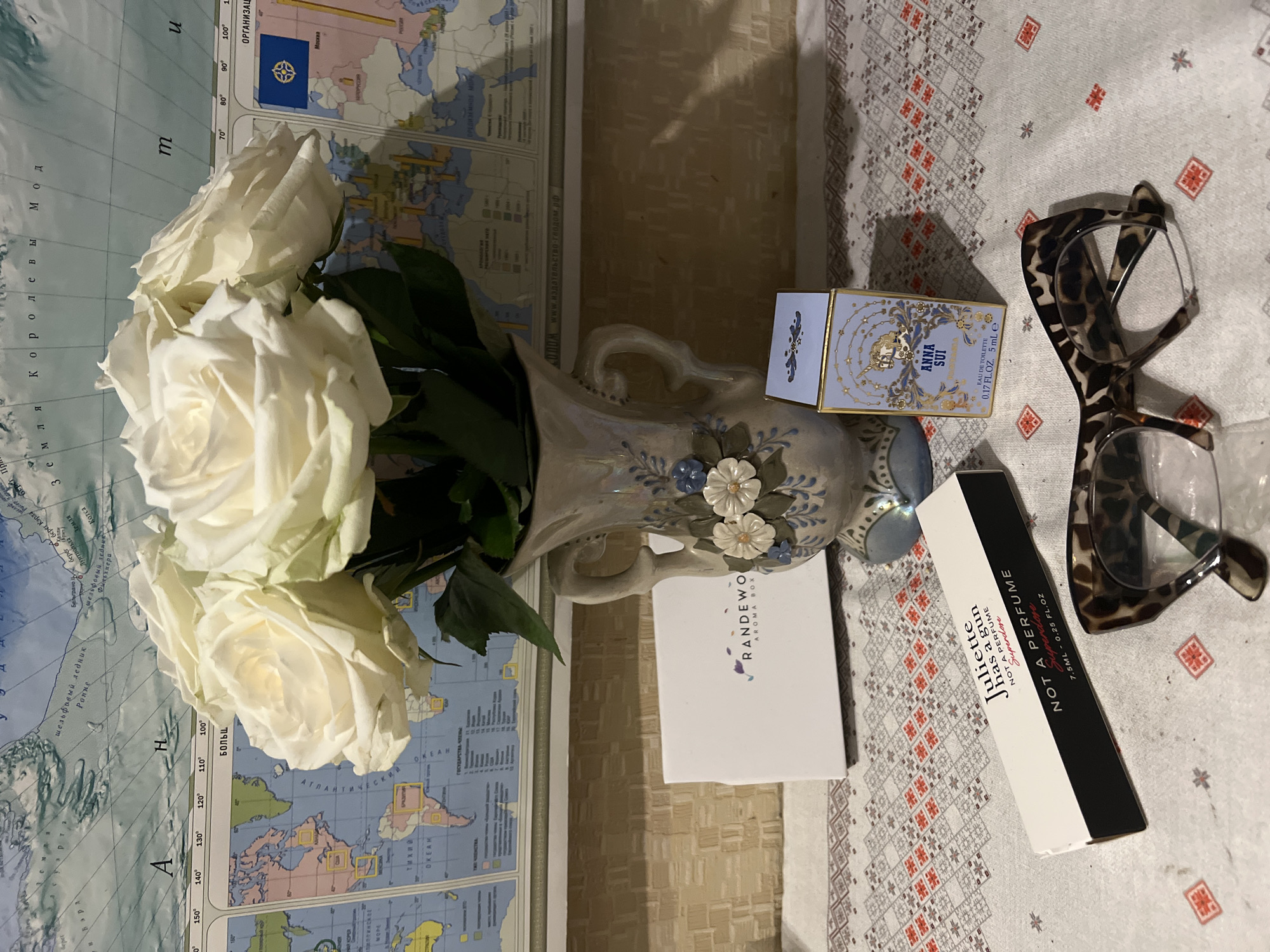 The image depicts a still life arrangement on a surface with a geometric pattern and a map backdrop. In the foreground, there is a vase filled with white roses. Next to the vase are several perfume boxes, namely "RANDEWO Aroma Box", "Juliette has a gun NOT A PERFUME Superdose", and "ANNA SUI Eau de Toilette". A pair of leopard-print eyeglasses are also visible in the arrangement. The map backdrop contains Russian text and depicts a geographic region. The image does not contain enough information to identify the city of Sochi, Russia.