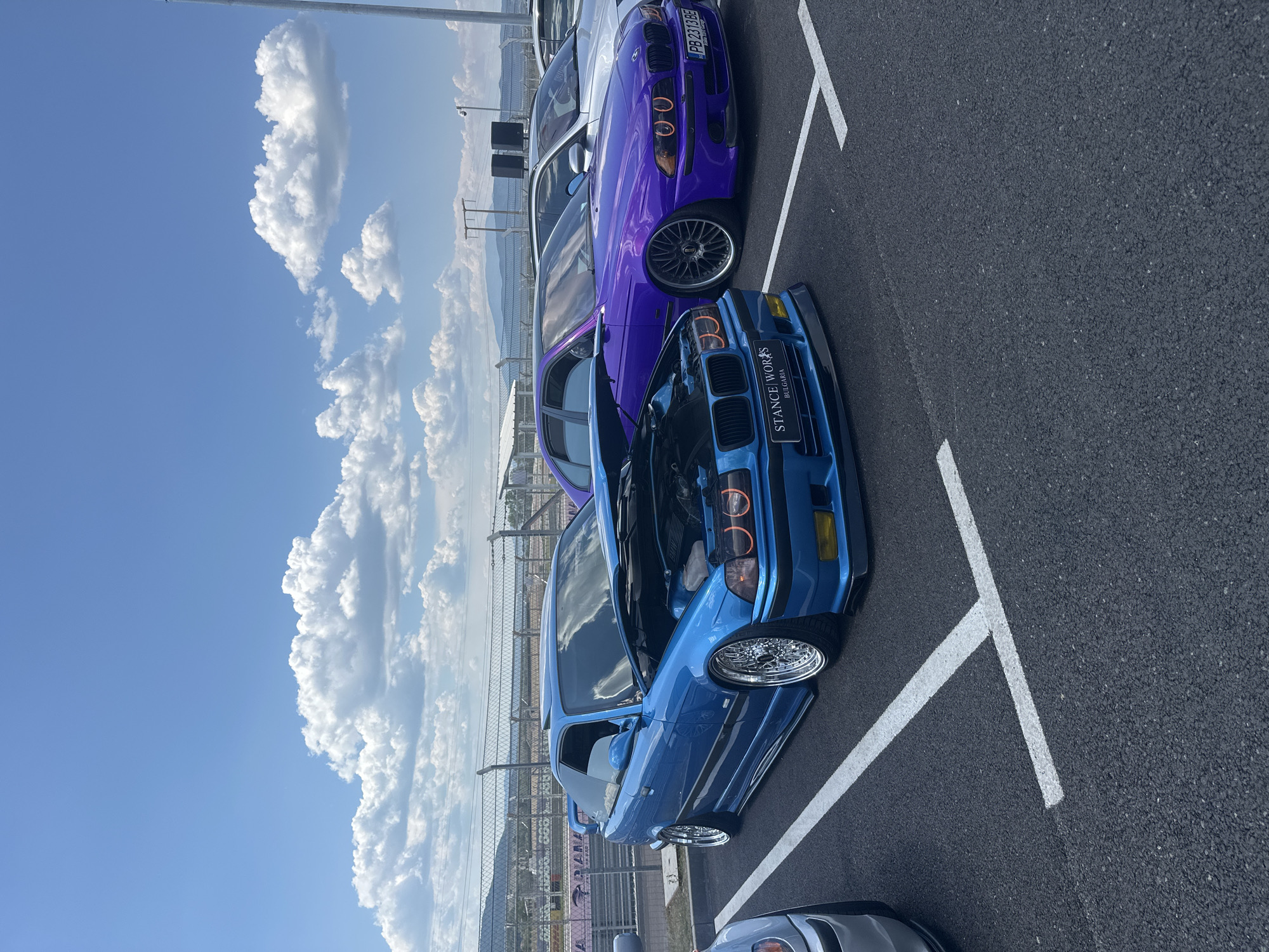 On a sunny day at the Race 77 circuit in Dospey, Bulgaria, a row of modified cars are parked on asphalt. The foreground features a bright blue BMW E36 coupe with its hood open, showcasing its engine. The car is lowered, with chrome rims and orange-accented headlights. To its right, a vibrant purple BMW E39 sedan is parked, also lowered and featuring dark rims and orange-accented headlights. Behind these vehicles, a silver BMW sedan is visible. The background includes a racetrack fence, grandstands, and distant mountains under a blue sky with scattered white clouds. The overall scene depicts a car gathering or show at a motorsport venue.